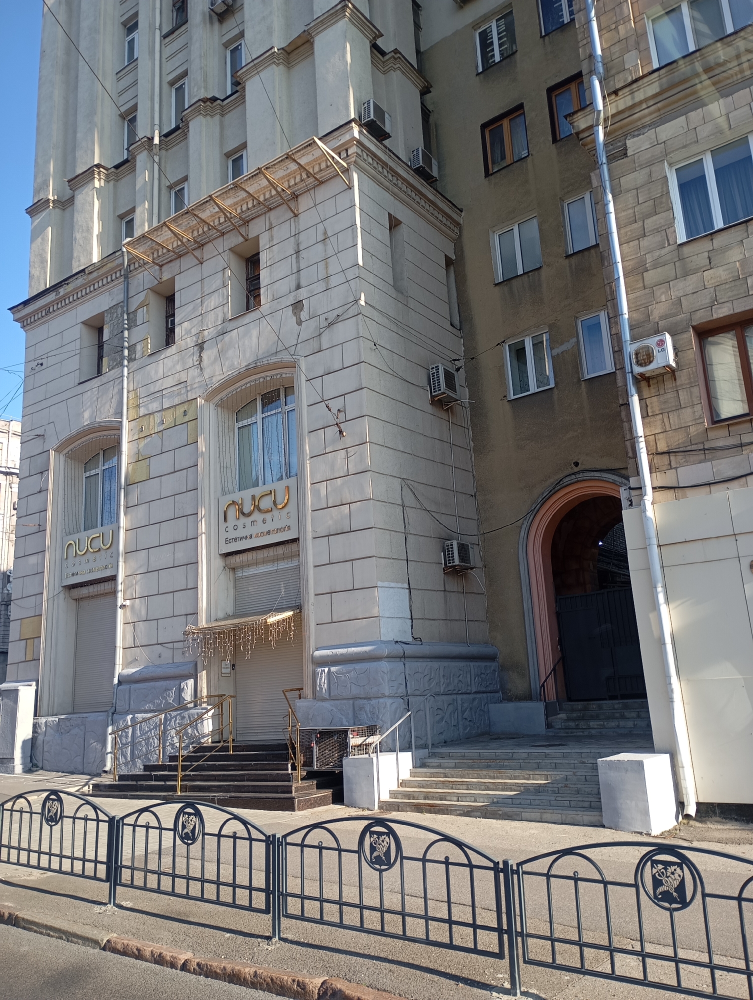 The image shows the facade of a building with a mix of architectural styles. The main building in the center is a light-colored stone structure with decorative elements and several windows. On this building, two prominent signs with the text "NUCLU COSMETIC" and "Eстетична косметологія" are visible above arched windows. To the right of this building, a narrower, darker facade is visible, featuring several windows and air conditioning units. This section also has an arched entryway. The buildings are bordered by a dark metal fence in the foreground, and a street with a sidewalk. The sky is clear and blue. The scene is located in Kharkiv, Ukraine, specifically on Sumska Street.