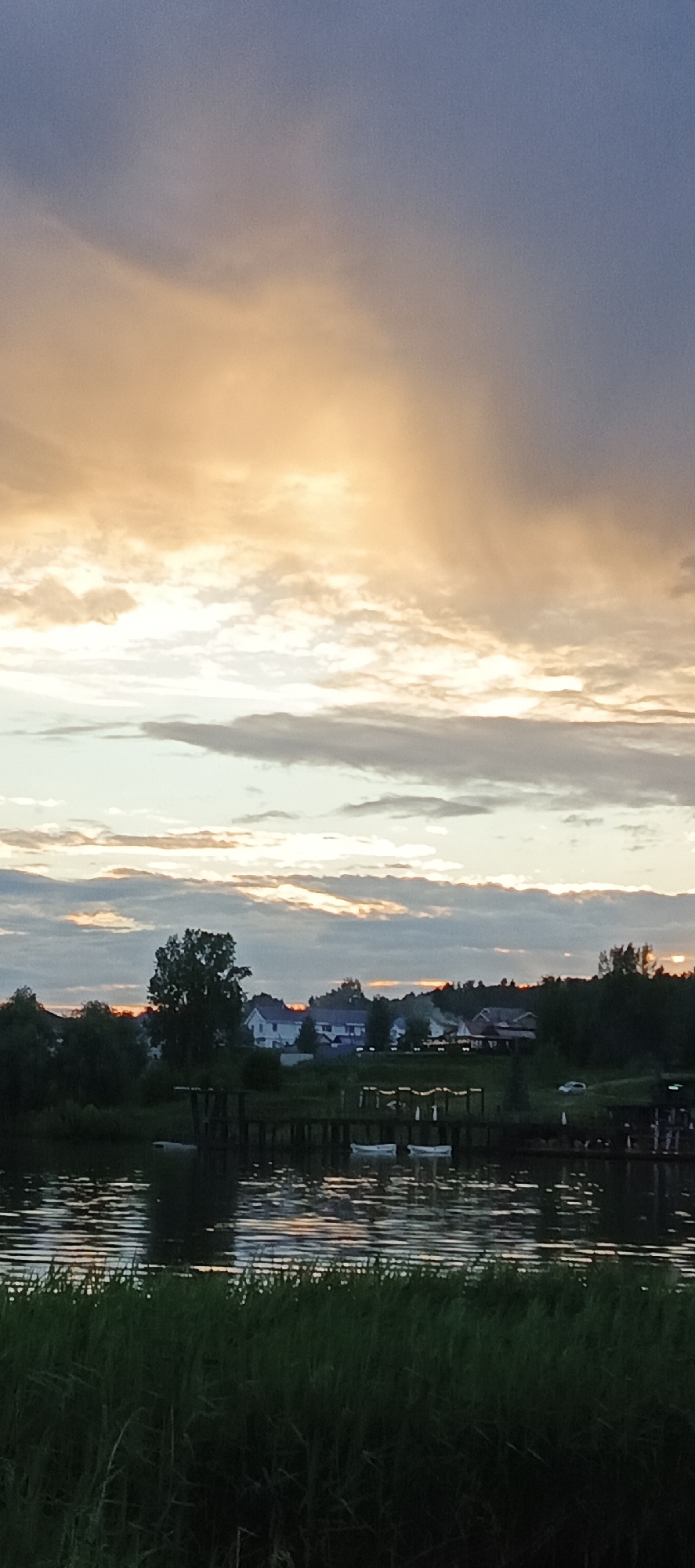 The image depicts a tranquil evening scene along a body of water, possibly a river or lake, with a Votkinsk, Russia, setting implied by the context of the original prompt. The sky is filled with dramatic clouds, transitioning from a warm, golden hue near the horizon to a soft blue overhead, suggesting either sunrise or sunset.
In the middle ground, a low-lying shoreline is visible, characterized by dense green trees and vegetation. Several houses, typical of a suburban or rural landscape, are scattered amongst the trees. A prominent dark green tree stands out against the lighter background.
In the foreground, the water's surface is rippled, reflecting the colors of the sky and the surrounding elements. Two small, white boats are moored near a wooden dock structure in the water, adding a sense of human presence and activity to the otherwise natural landscape. The water's edge is lined with tall, dark green grass or reeds. A small, white car is parked on the land near the houses on the right side of the image. The overall atmosphere is serene and picturesque.