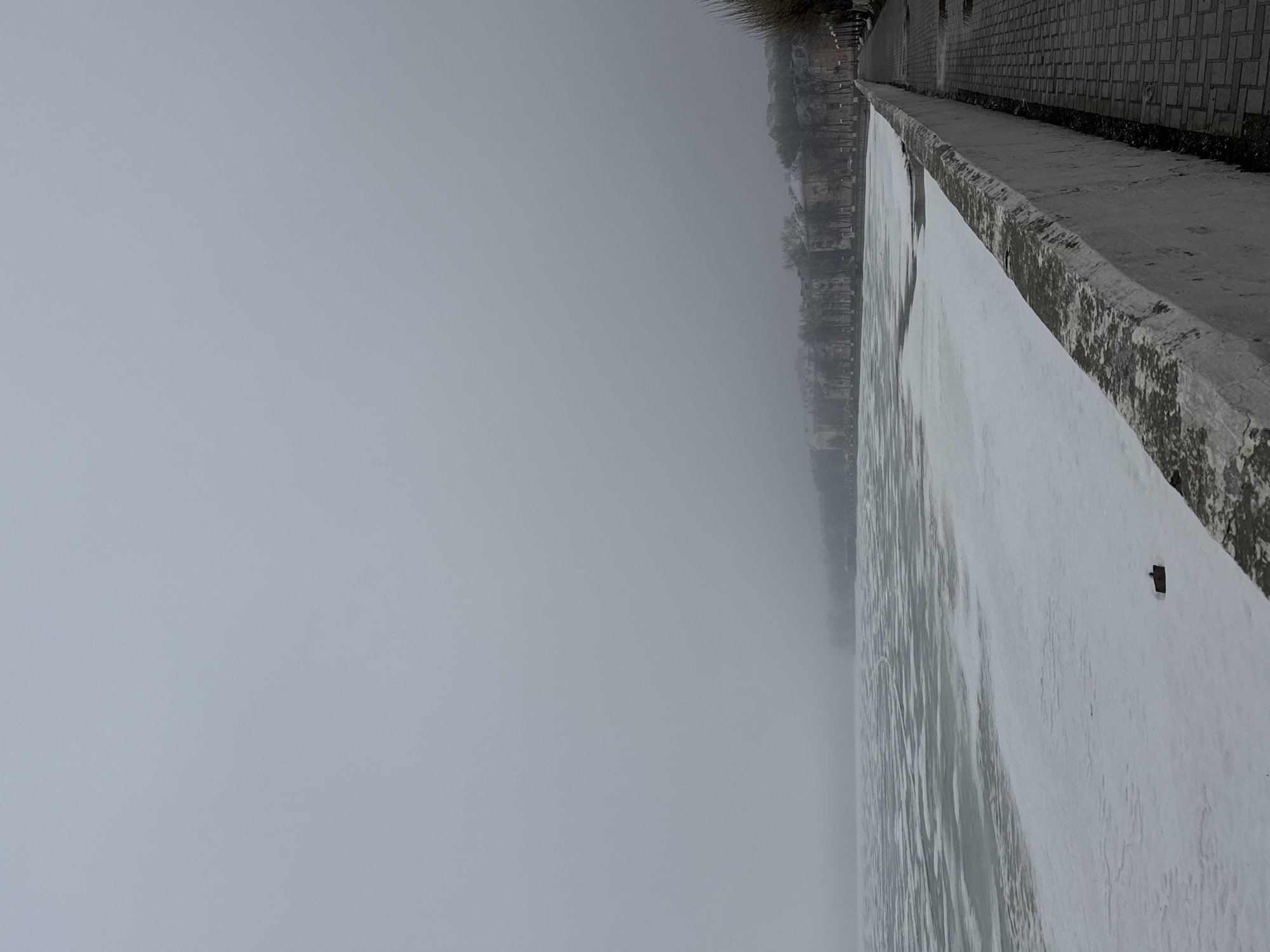 The image depicts a foggy scene in Votkinsk, Russia, alongside a river embankment. A concrete wall runs along the right side of the frame, with a paved walkway behind it. Buildings are visible through the fog in the background. The foreground shows what appears to be frozen water or ice covering the river surface.