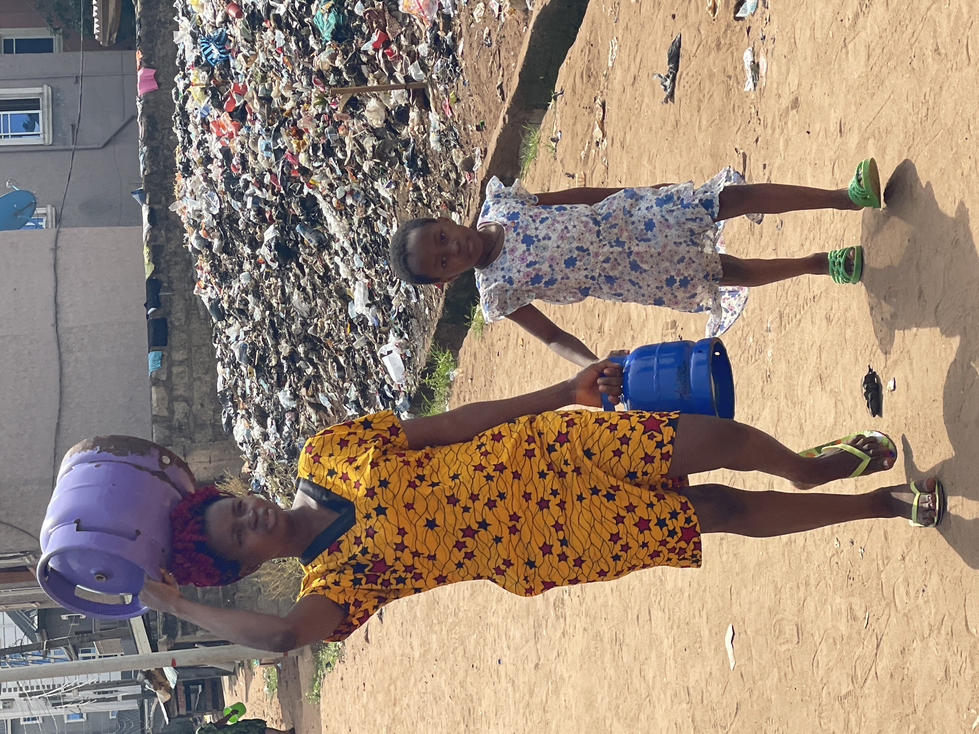 The scene is set in Nkpor, Nigeria. An adult woman in a yellow star-patterned dress carries a purple gas cylinder on her head. A young girl in a floral dress is holding a blue canister. Both are on a dirt ground. In the background, a large pile of garbage is visible. A building is partially seen on the left side.
