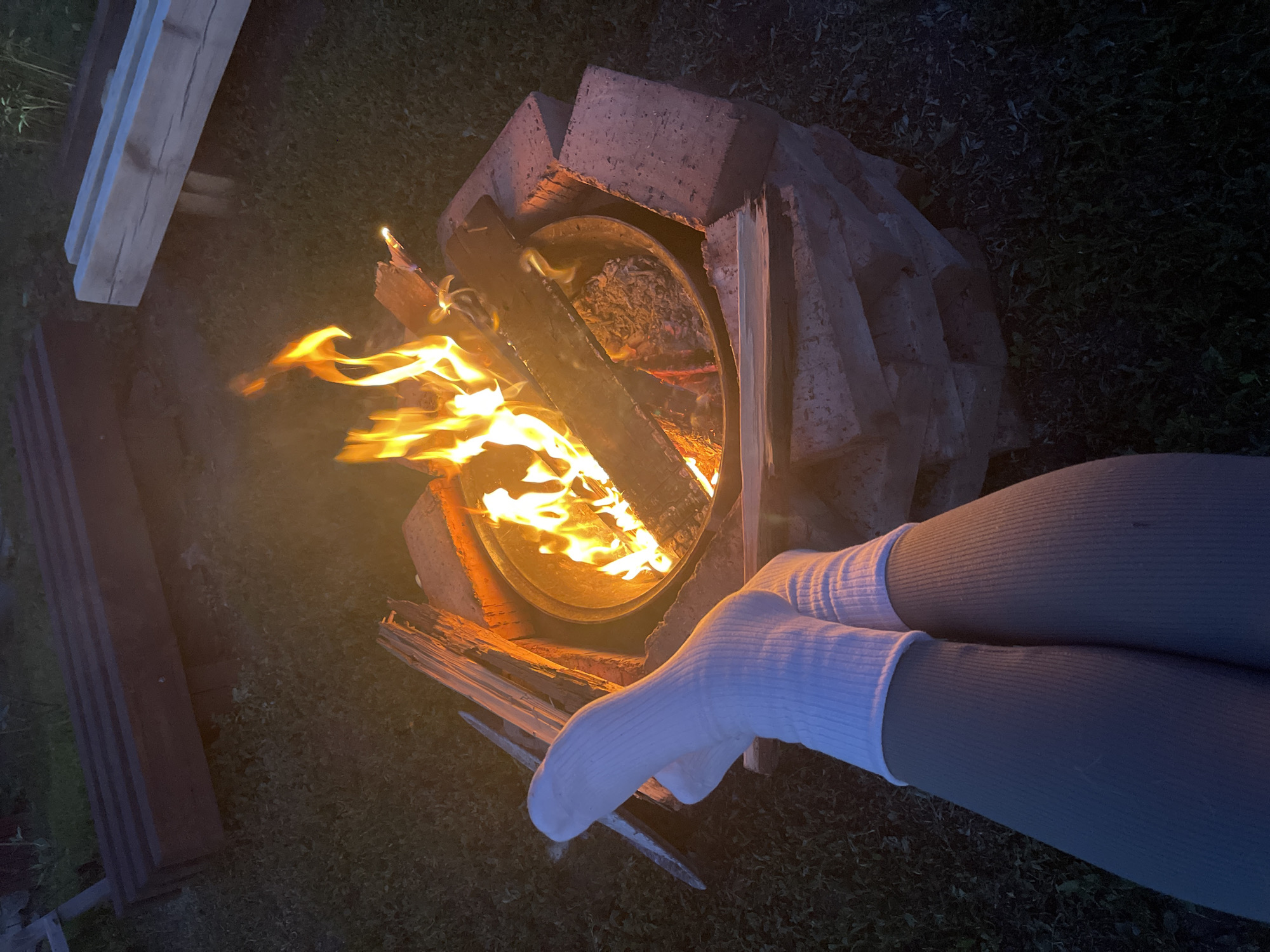 In an outdoor setting with a dark background, a person's legs, clad in white socks and grey leggings, are crossed in the foreground, facing a brick fire pit. The fire pit contains a roaring fire with flames reaching upwards. Scattered around the fire pit are pieces of firewood and some ash. In the background, there are two wooden structures, possibly benches or tables, one appearing to be made of dark wood and the other of lighter wood. The ground around the fire pit is covered in grass and scattered leaves. The overall scene suggests a cozy evening gathering around a campfire.