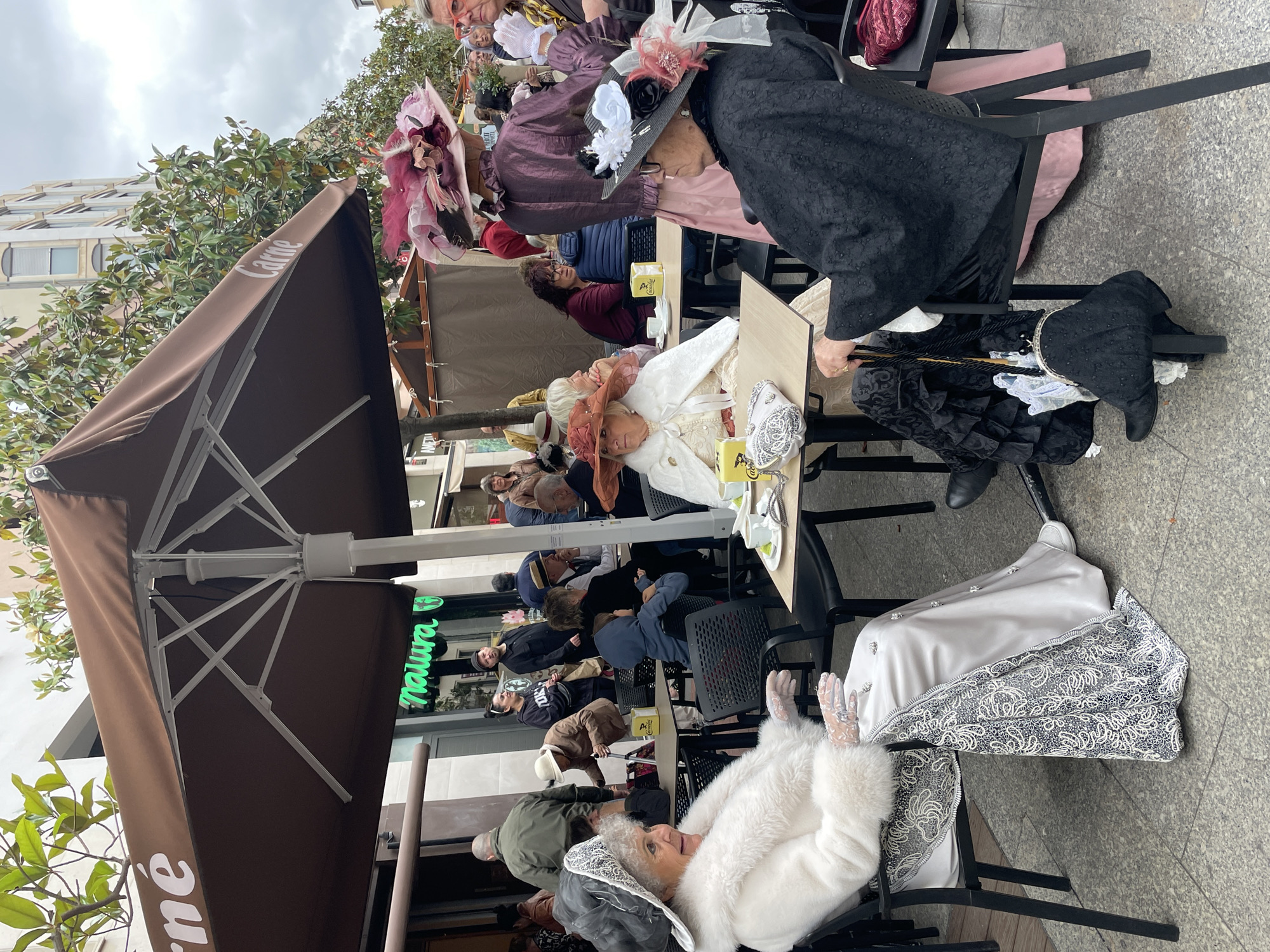 The image shows a group of elderly women dressed in period costumes, sitting at tables outside a cafe in Terrassa, Spain. The women are wearing elaborate hats and dresses. Some of the women are interacting with each other. There are other people at the tables that are not in costume. The cafe has brown umbrellas with the word "Carne" printed on them. There are trees and buildings in the background. The weather appears overcast.