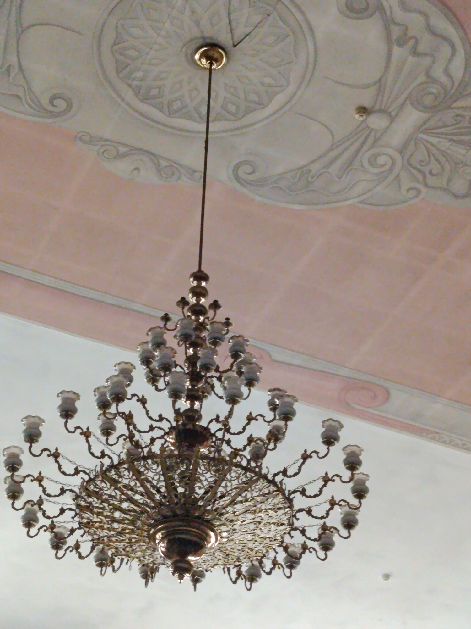 The image shows a chandelier hanging from a decorated ceiling in Moscow, Russia. The chandelier is ornate, made of metal with multiple arms holding glass lamps. The ceiling features intricate patterns and is painted in soft colors, with decorative molding. The overall impression is of an interior space, possibly a historical building or a grand hall.
