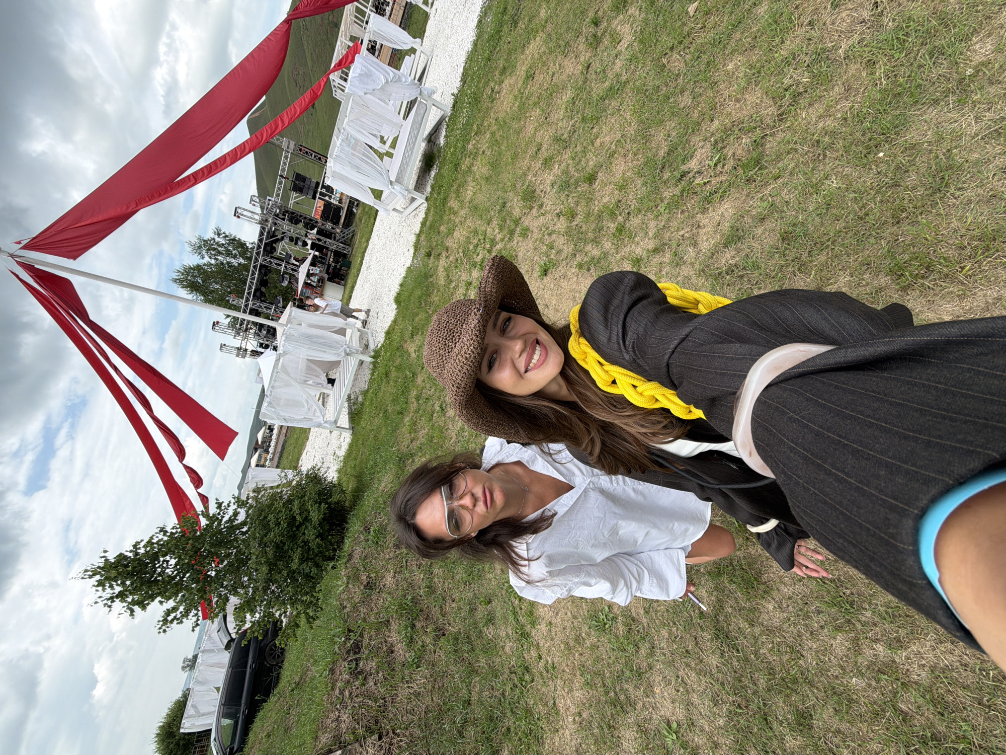 Two women are taking a selfie outdoors on a cloudy day. They are dressed casually, with one wearing a white shirt and glasses, and the other sporting a brown hat, a dark pinstripe jacket, and a bright yellow scarf. The woman in the hat is smiling at the camera, while the other woman looks more serious.
In the background, a large stage setup with multiple speaker systems and lighting rigs is visible, suggesting an outdoor event or concert. Red fabric banners are suspended from a large white pole, adding a festive touch to the scene. Several white canopied daybeds are also set up on the grassy area, indicating a comfortable lounging space for attendees. The overall setting appears to be an open field, possibly a park or an event venue, with rolling hills in the distance. The lighting suggests it is daytime.