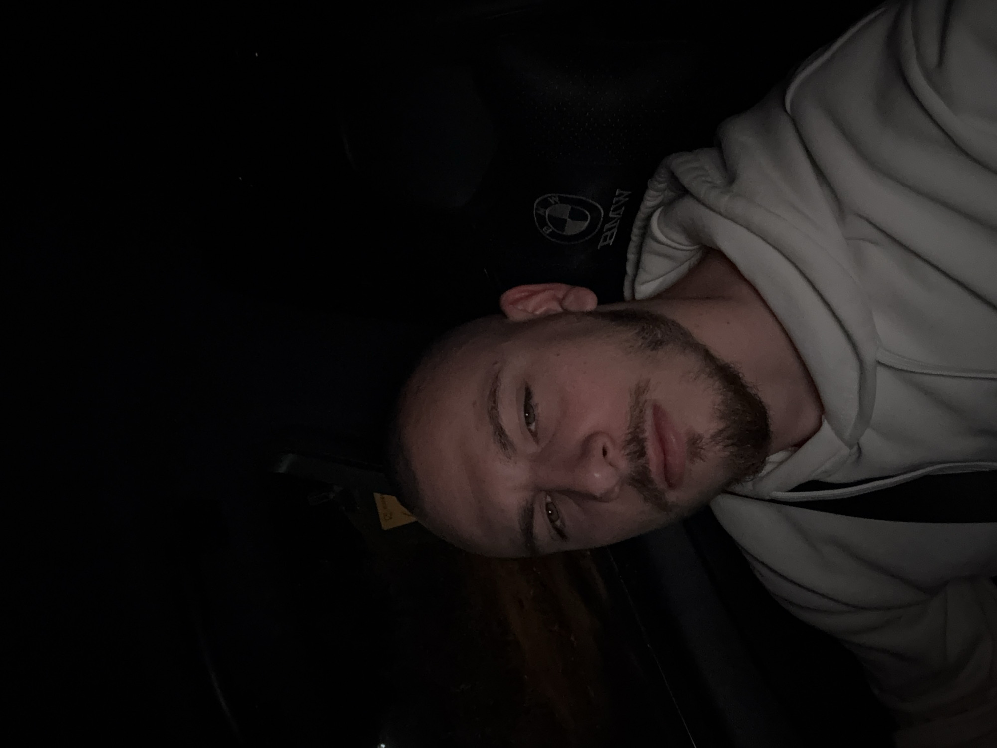 The image shows a man in the interior of a car, possibly a BMW, as indicated by the logo on the headrest. The man has a short haircut and a beard, wearing a white hoodie. The environment is dark, suggesting night. It is impossible to determine the location in Stara Zagora, Bulgaria based on this image.