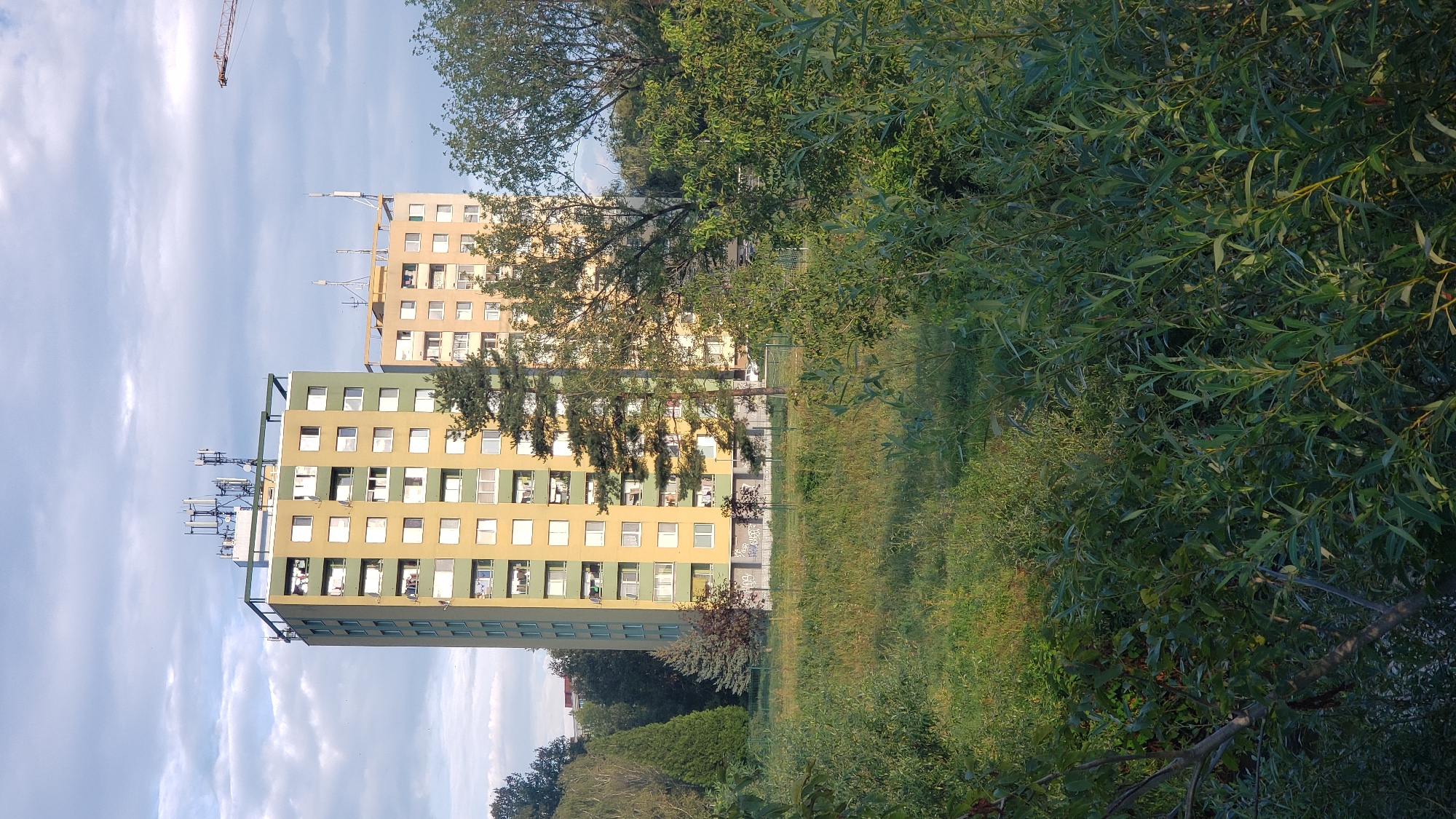 The image depicts two tall apartment buildings, one predominantly yellow with green accents and the other a lighter yellow, situated in the city of Prešov, Slovakia. The buildings have multiple floors with visible windows, and the taller yellow and green building on the left features numerous antennas on its roof, suggesting a telecommunications hub. A construction crane is partially visible in the upper right portion of the frame. The buildings are surrounded by a variety of trees and dense vegetation in the foreground, with a grassy field separating the trees from the apartment blocks. The sky is partly cloudy, with a soft, diffused light indicating it is likely late afternoon or early evening. The scene appears to be a residential area, likely a housing estate, with a mix of urban structures and natural elements.