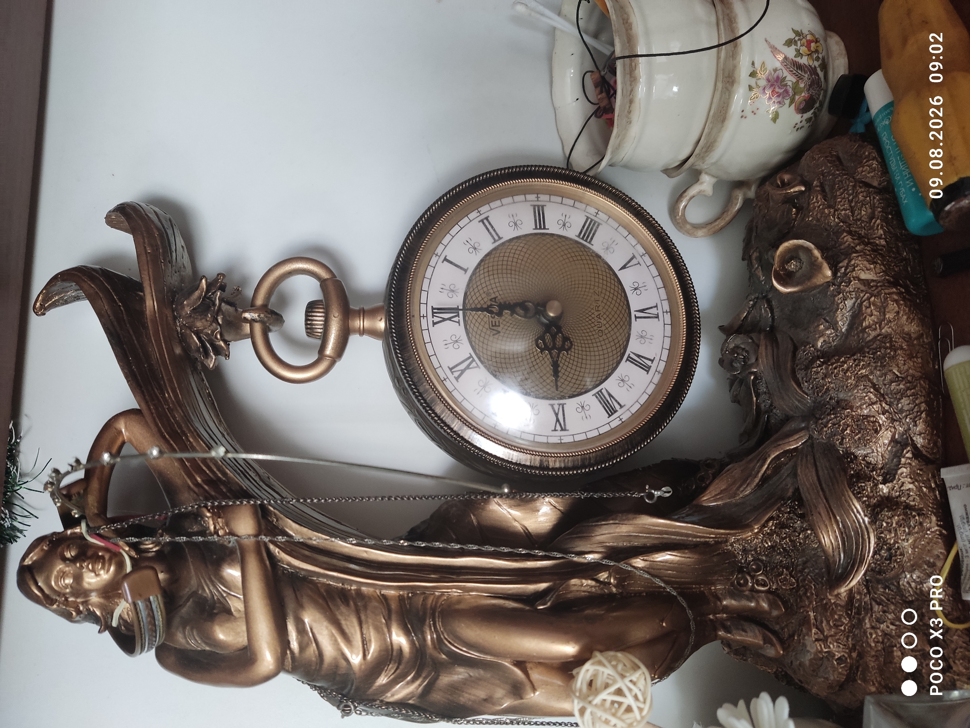 The image depicts a still life arrangement on a shelf. Dominating the left side is a bronze-colored statue of a woman holding a laurel wreath, from which a pocket watch is suspended. The watch has a Roman numeral face and appears to be a Vedia brand quartz timepiece. To the right of the watch are two stacked ceramic cups, the bottom one decorated with a floral and bird motif. The background is a plain white wall. In the foreground, various objects are visible including what appears to be a tube of medicine or cosmetic, and a portion of a wooden surface. The lighting suggests an indoor environment, possibly a home or a display shelf. The timestamp in the lower right corner indicates "09.08.2026 09:02", and the lower left corner shows "POCO X3 PRO". There are no people present in the image. This scene does not contain any information that can be used to determine its location in the city of Cherniakhiv, Ukraine.
