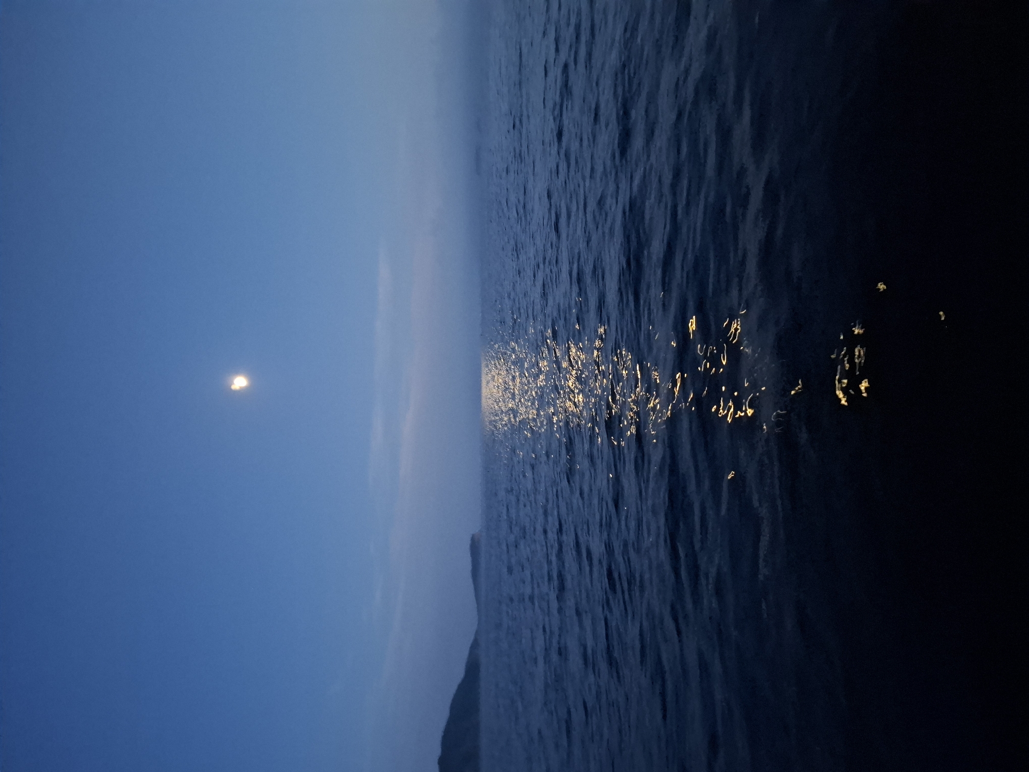 The image depicts a body of water under a twilight sky, with a moon visible in the upper portion of the frame. A reflection of the moon shimmers on the water's surface, creating a path of light. In the distance, a hazy shoreline with landmasses is visible, suggesting a coastal or island location. The overall scene conveys a sense of calm and natural beauty. The location is Putyatin Island, Russia, known for its scenic landscapes and marine environment. There are no people or specific objects of human construction visible in the image, focusing solely on the natural elements of the seascape.