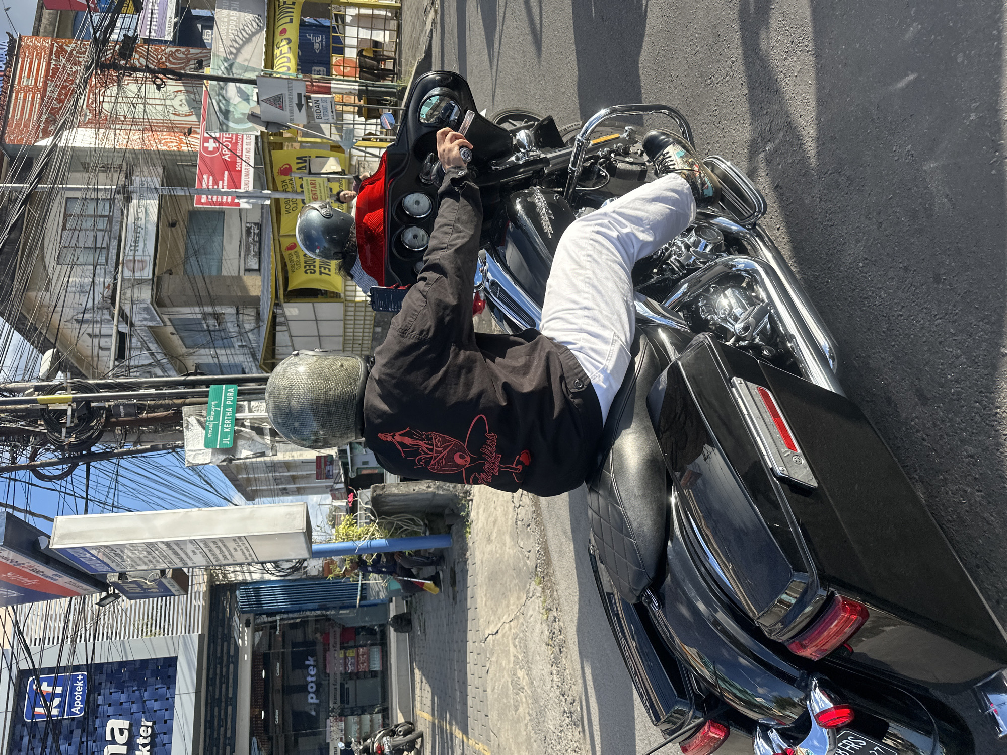 The image captures a street scene in Denpasar, Indonesia, focusing on a person riding a black Harley-Davidson motorcycle. The rider, seen from behind, wears a dark jacket with a red graphic print and white pants. Their head is covered by a silver helmet with a textured surface. The motorcycle is equipped with saddlebags and chrome accents. In the background, there are buildings with signs indicating businesses, including an "Apotek" (pharmacy) with the "Apotek+" logo. Overhead, a complex network of electrical wires is visible against a blue sky. A green street sign with text in a local script and "JL. KERTHA PURA" is also present, indicating the location within Denpasar. The overall environment suggests a typical urban street with commercial establishments and infrastructure.