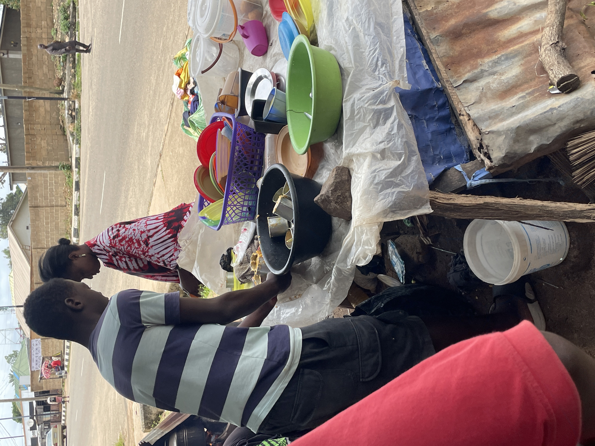 In the city of Jos, Nigeria, a street vendor is captured interacting with a customer on an overcast day. The vendor, a young man wearing a purple and light blue striped t-shirt and dark cargo shorts, is bent over, his hands near a collection of plastic containers and dishes spread out on a white plastic sheeting. Beside him, a woman in a vibrant red and white patterned traditional attire is also engaged in the transaction, her head bowed slightly towards the vendor. The background shows a dusty road with a few distant buildings and trees, suggesting an outdoor market or roadside stall. The scene is one of everyday commerce, with various domestic items, including buckets, bowls, and plates, laid out for sale. The ground is a mix of dirt and scattered debris, with a corrugated iron structure visible on the right side of the frame, contributing to the rustic, informal setting.