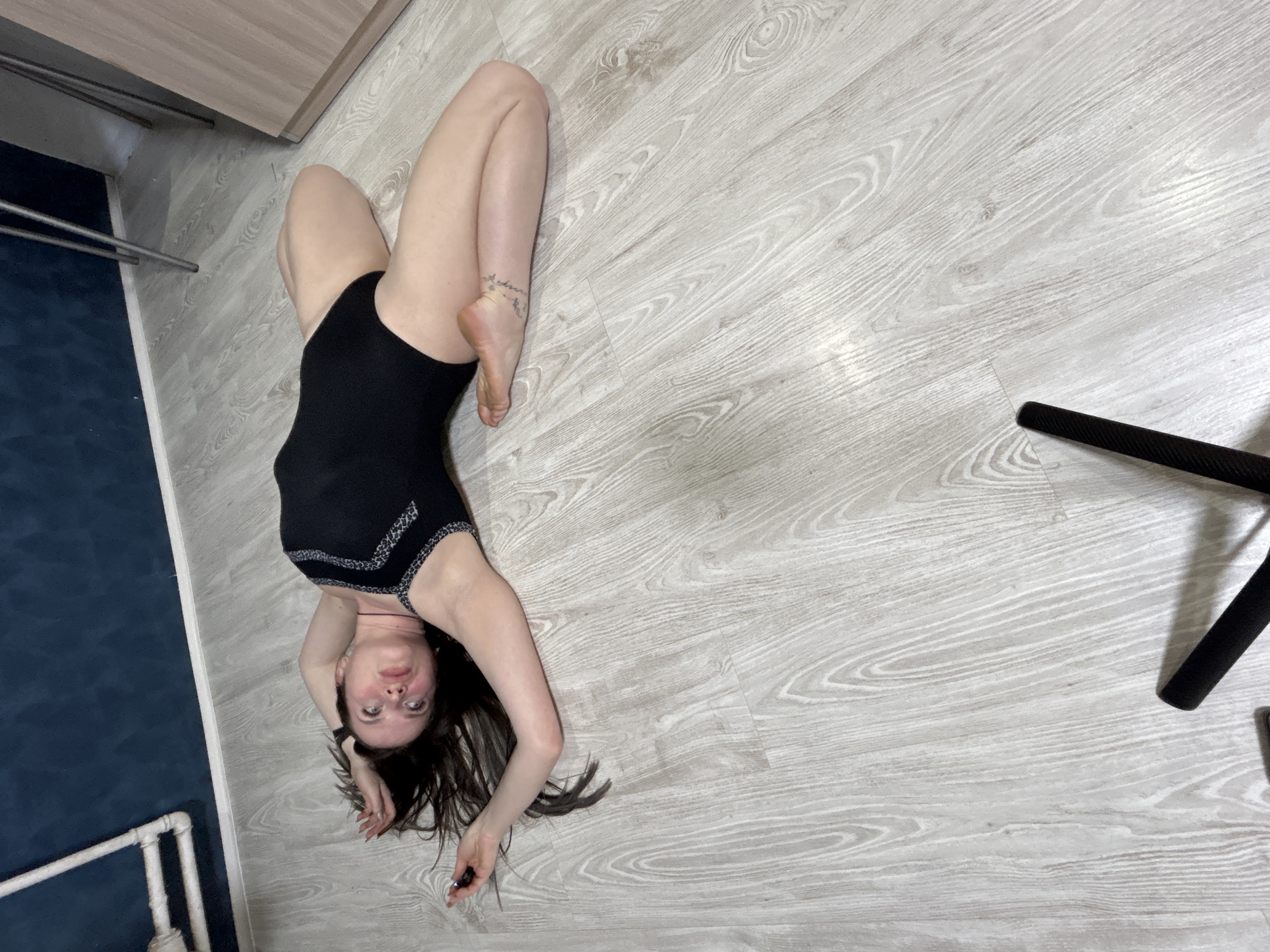 A woman is depicted lying on her back on a light wood-effect floor in an indoor setting, likely a room in Rusalevka, Russia. Her head is positioned towards the bottom of the frame, with her dark hair splayed around her head. She looks directly up at the camera with a neutral expression. She wears a black one-piece garment, possibly a leotard or swimsuit, which has a distinctive silver or light-colored patterned trim along the chest and waist. Her legs are bent, with her feet positioned close to her head, demonstrating flexibility. A small, illegible cursive tattoo is visible on her right ankle, and she wears a watch on her left wrist.
The floor is a light-colored laminate or vinyl with a wood grain pattern. To the left, a dark blue or grey wall meets the floor, delineated by a white trim or baseboard. Above this dark wall, a light wood-toned cabinet or cupboard is visible. Further to the left, against the dark wall, are some metal bars, possibly part of a drying rack or furniture. On the right side of the frame, a black object, likely a tripod leg, is partially visible on the floor. The lighting is even, suggesting an indoor environment without harsh shadows.