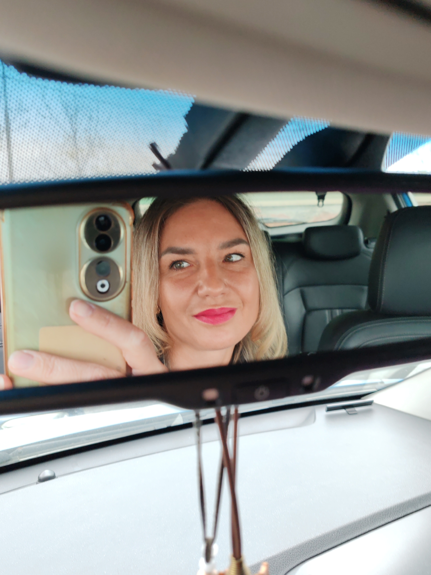 A fair-skinned woman with light brown hair is taking a selfie in a car using her smartphone. She is visible in the rearview mirror, her left eye looking slightly to the side. She is wearing bright pink lipstick. The smartphone has two prominent camera lenses on its back. The back seats of the car, upholstered in black leather, are visible in the mirror's reflection. The car's dashboard is light gray. There is a hanging ornament in front of the rearview mirror. The top of the windshield shows a blue, dotted pattern, suggesting a sun visor strip. The background outside the car is blurred. The location of the photo cannot be determined to be Petropavlovsk-Kamchatsky, Russia from the image alone.