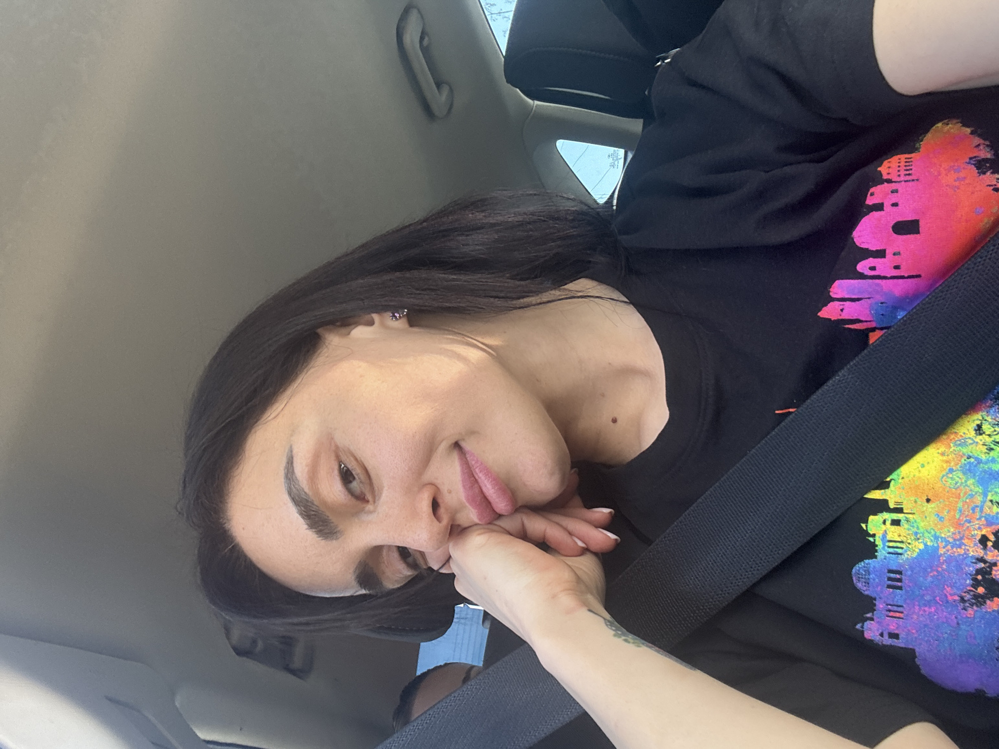 A person is in a car with a seatbelt on, wearing a black T-shirt with a colorful graphic. The person is leaning their face on their hand. The environment appears to be the interior of a vehicle. There's no indication of Tbilisi, Georgia.