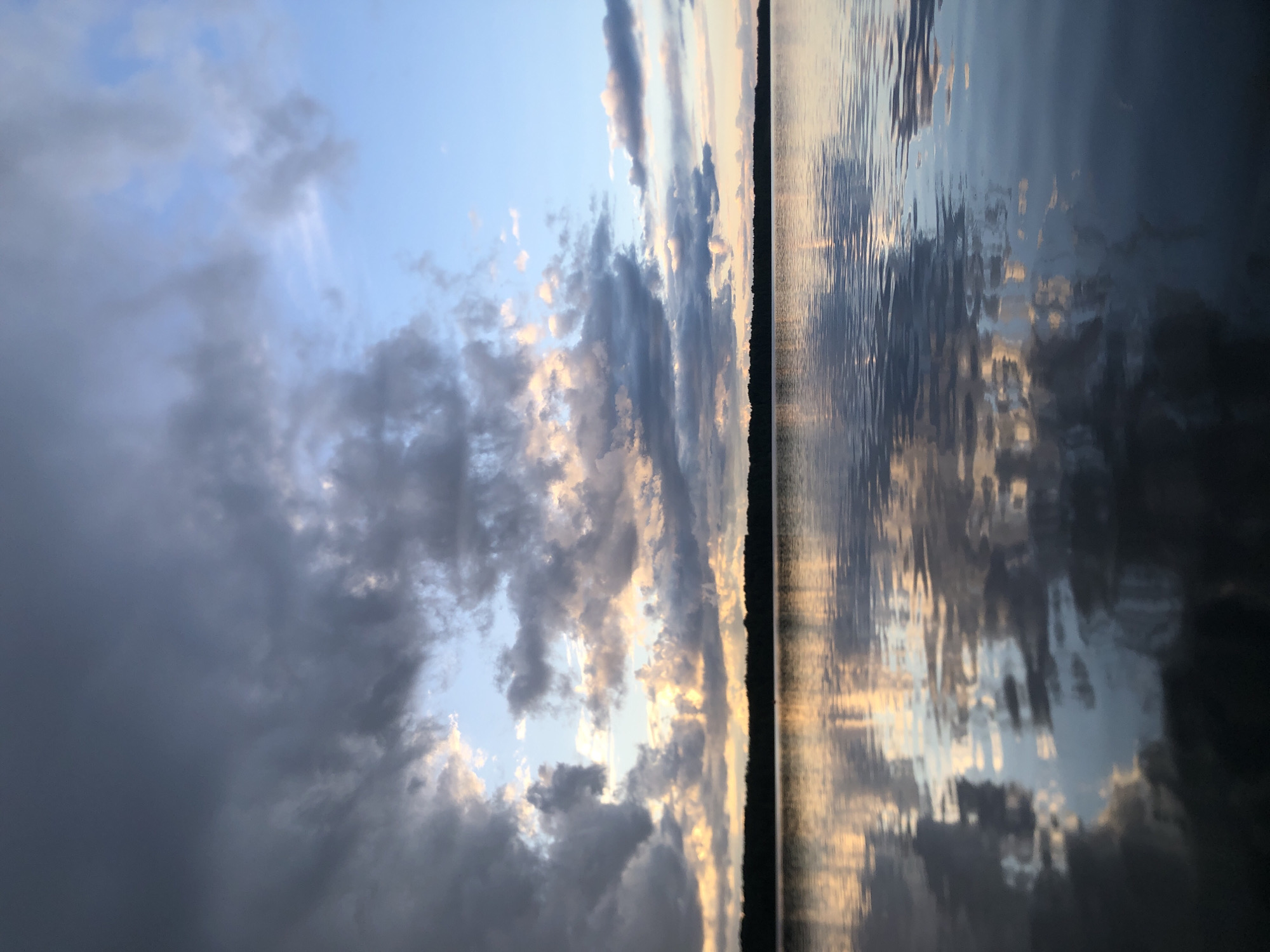 This media file depicts a serene natural scene with a prominent body of water reflecting a dramatic sky. The image is oriented vertically, with the sky occupying the left half and the water the right.
The sky is filled with a mixture of dark, moody storm clouds and patches of brighter, illuminated clouds, suggesting a transition in weather or the approaching end of the day. Sunlight, possibly from a setting sun, breaks through in some areas, casting warm golden hues on the edges of the clouds. The overall impression of the sky is dynamic and atmospheric.
The water surface is rippled, creating abstract patterns that mirror the sky's colors and cloud formations. These reflections are fragmented and distorted by the movement of the water, adding a sense of depth and texture to the scene. A thin, dark line runs vertically along the boundary between the sky and water, likely representing a distant treeline or the edge of land.
The time of day appears to be late afternoon or early evening, given the quality of light and the cloud formations. The weather seems unsettled, with the presence of dark clouds. The location could be a lake, a wide river, or a coastal area, given the vastness of the water and sky. The emotion conveyed is one of peacefulness, awe, and perhaps a touch of melancholy due to the stormy elements in the sky. There is no visible text in the image.