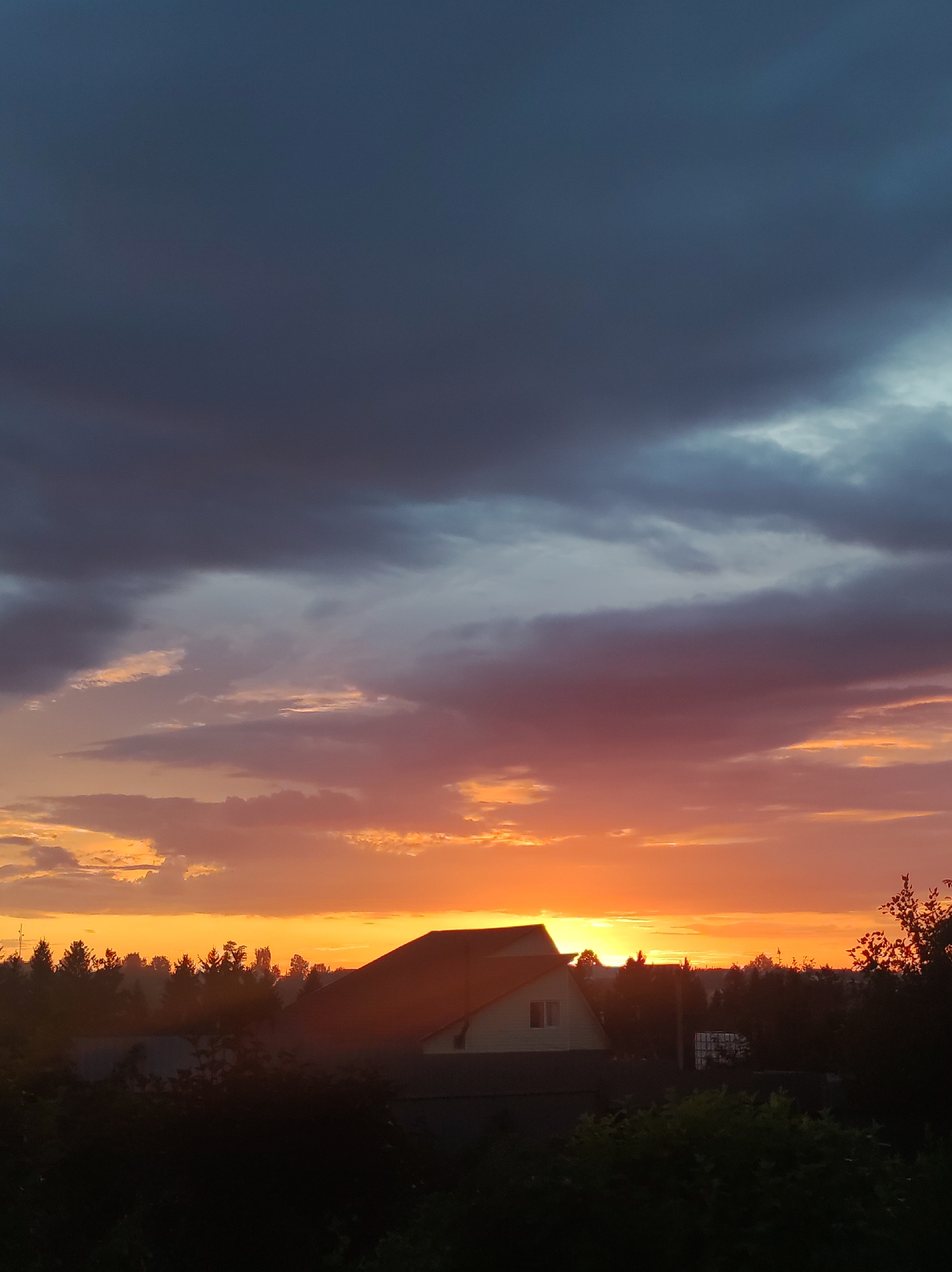 The image shows a sunset over the city of Foky, Russia. The sky is filled with dramatic clouds, ranging from dark gray at the top to vibrant oranges and pinks near the horizon. Silhouetted against the colorful sky are the rooftops of several houses, including a prominent house with a light-colored facade and a dark roof in the foreground. A line of trees and other foliage forms a dark band across the lower portion of the image, obscuring the ground and the lower parts of the buildings. The overall impression is one of a serene, yet visually striking, evening scene.