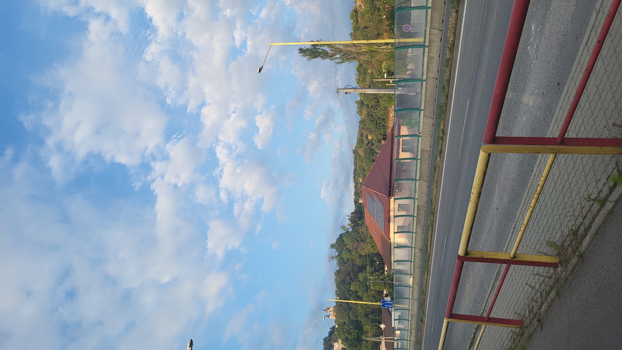 The image depicts a roadside scene with a highway in the foreground and a suburban landscape in the background under a partly cloudy sky. The highway, with visible lane markings, is bordered by a red and yellow metal railing and a paved pedestrian walkway. Beyond the highway, a green sound barrier is present, followed by a house with a red tile roof and solar panels. Further in the distance, a hill covered with trees is visible, with a church or religious building topped with domes on the left side of the hill. A tall, slender tree stands near a lamppost on the right side of the frame. The overall scene suggests a transition between urban and natural environments. Based on the visual elements, particularly the style of architecture and the presence of the specific religious building, this scene is likely located in Prešov, Slovakia, possibly on a road leading into or out of the city.