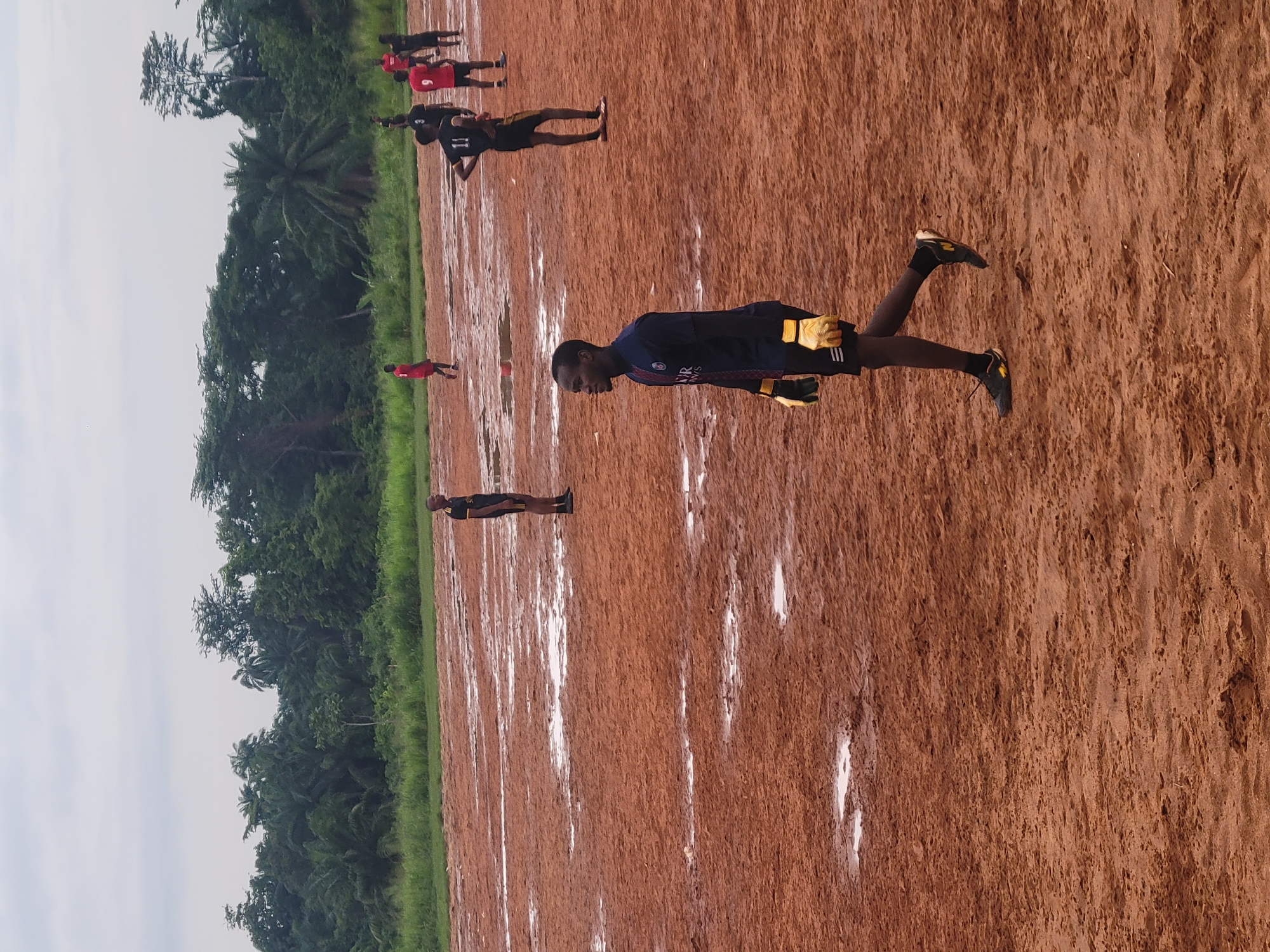 This media file shows a group of young men on a dirt field, likely a sports field, in Egbagu, Nigeria. The field is marked with white lines, suggesting it is used for races or other athletic events. The ground is a reddish-brown, indicative of the soil type in the region. In the background, there is dense green vegetation, including palm trees, characteristic of a tropical environment. The sky appears overcast, suggesting a cloudy or possibly rainy day.
Several individuals are visible, mostly wearing athletic attire. One person in the foreground is wearing a dark blue shirt with yellow trim and yellow gloves, possibly a goalkeeper or a participant in a specific sport. Another person further back is wearing a red shirt and black shorts, while others are in dark athletic wear. The people are in various poses, some appearing to be running or preparing to run, while others are standing. This suggests an athletic training session or a competition is taking place. The overall atmosphere seems to be one of activity and focus. No discernible text is visible on the clothing or surroundings.