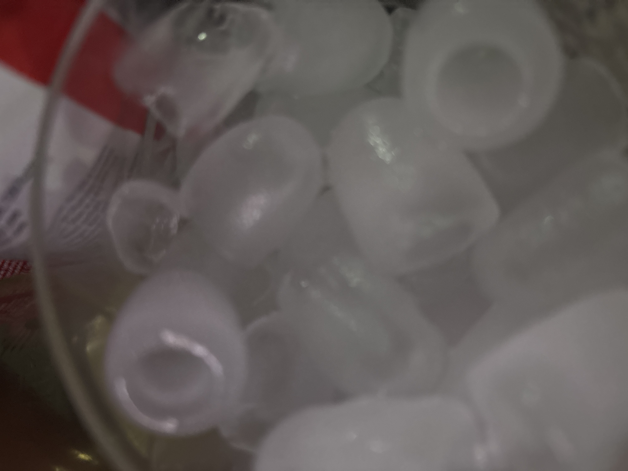 The image shows a close-up, out-of-focus view of ice cubes in a glass. The ice cubes are predominantly white and translucent, with some appearing cylindrical and others more irregular in shape. The lighting is soft and diffused, creating highlights and shadows on the surfaces of the ice. The background is a blur of muted colors, suggesting the presence of other objects or a dark environment. There are no visible signs of people, specific actions, or interactions. The scene is devoid of any environmental context that would indicate a location in Trudovets, Bulgaria, or any other specific geographic area.