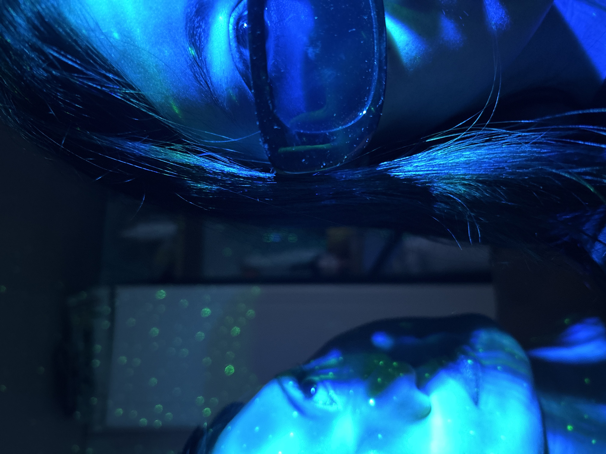 The image depicts two individuals within a darkly lit, atmospheric setting, bathed in intense blue light. The context provided indicates the location is Фоки, Russia. The upper subject's face is partially visible from an unusual angle, showing an eye, forehead, and dark hair, and they are wearing glasses that reflect the strong blue illumination. Their skin and hair are vividly highlighted by the neon-like blue glow. Below them, another person's face, specifically a cheek and chin, is also visible and similarly illuminated. Both subjects, as well as the dark background, are adorned with numerous small, bright green, circular light specks, which appear to be projected laser dots or a similar dynamic light effect. The setting is likely indoors, in a dark room or at night, creating a dreamlike or futuristic ambiance. The general activity seems to be an immersive experience with a light show or special lighting. The close proximity of the two individuals suggests a shared and intimate moment. No text is visible within the image.