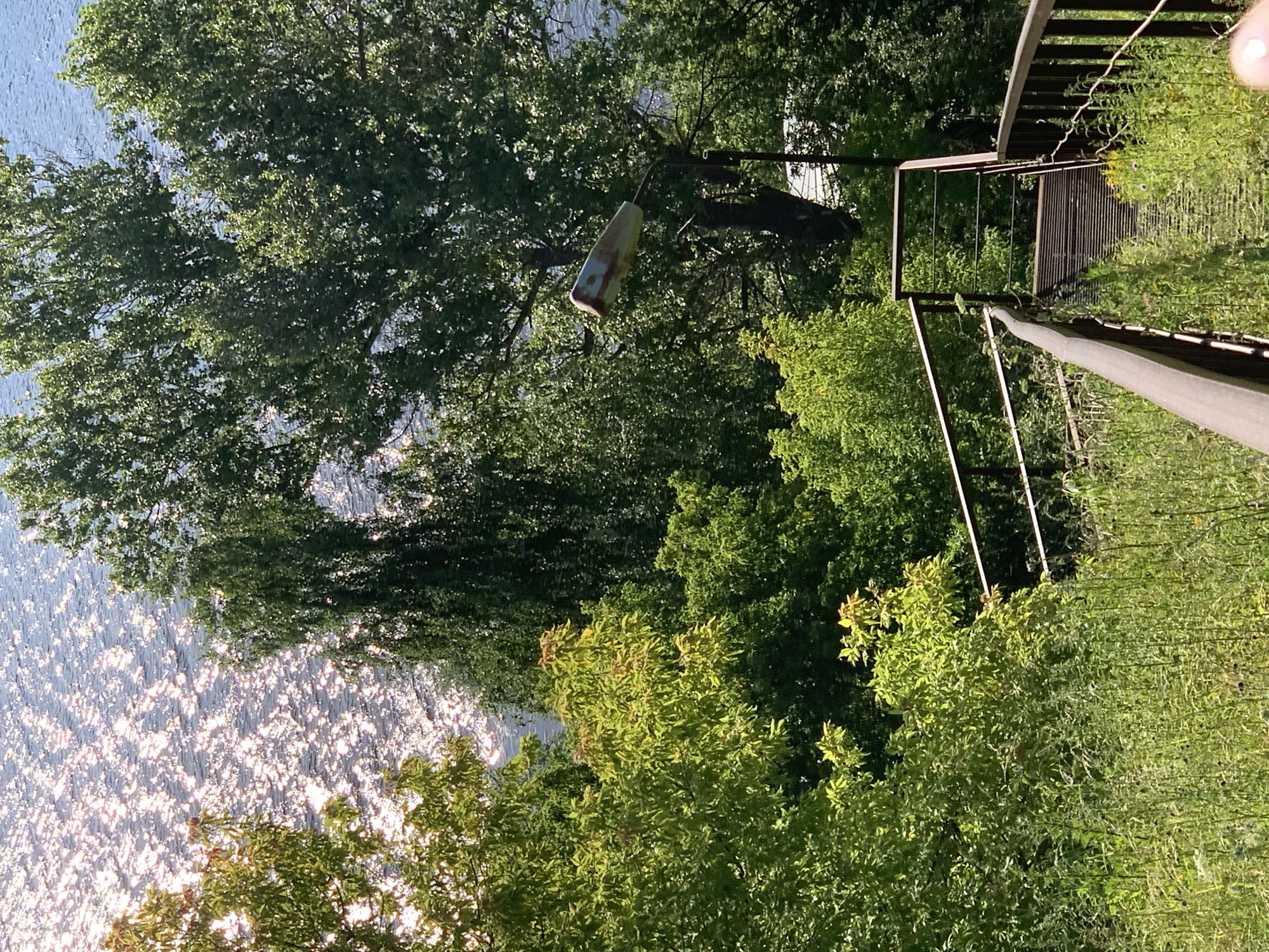This media file captures a sun-dappled scene, likely during the daytime with clear weather, in Pavlovo, Russia. The primary focus is a tranquil body of water, possibly a river or lake, reflecting the bright sunlight in a shimmering pattern. Lush green trees with dense foliage surround the water, creating a natural and serene environment.
A rustic wooden walkway or bridge with metal railings is visible, leading down towards the water. The structure appears weathered, suggesting it has been there for some time. Hanging from a branch of a tree is an old, somewhat worn-out buoy, its surface showing hints of faded colors. This object adds a touch of nostalgia and hints at past activity related to the water.
The overall impression is one of peacefulness and natural beauty, typical of a summer day in a rural or semi-rural setting. The light filtering through the leaves and the sparkling water contribute to a tranquil and inviting atmosphere. There is no visible text within the image.