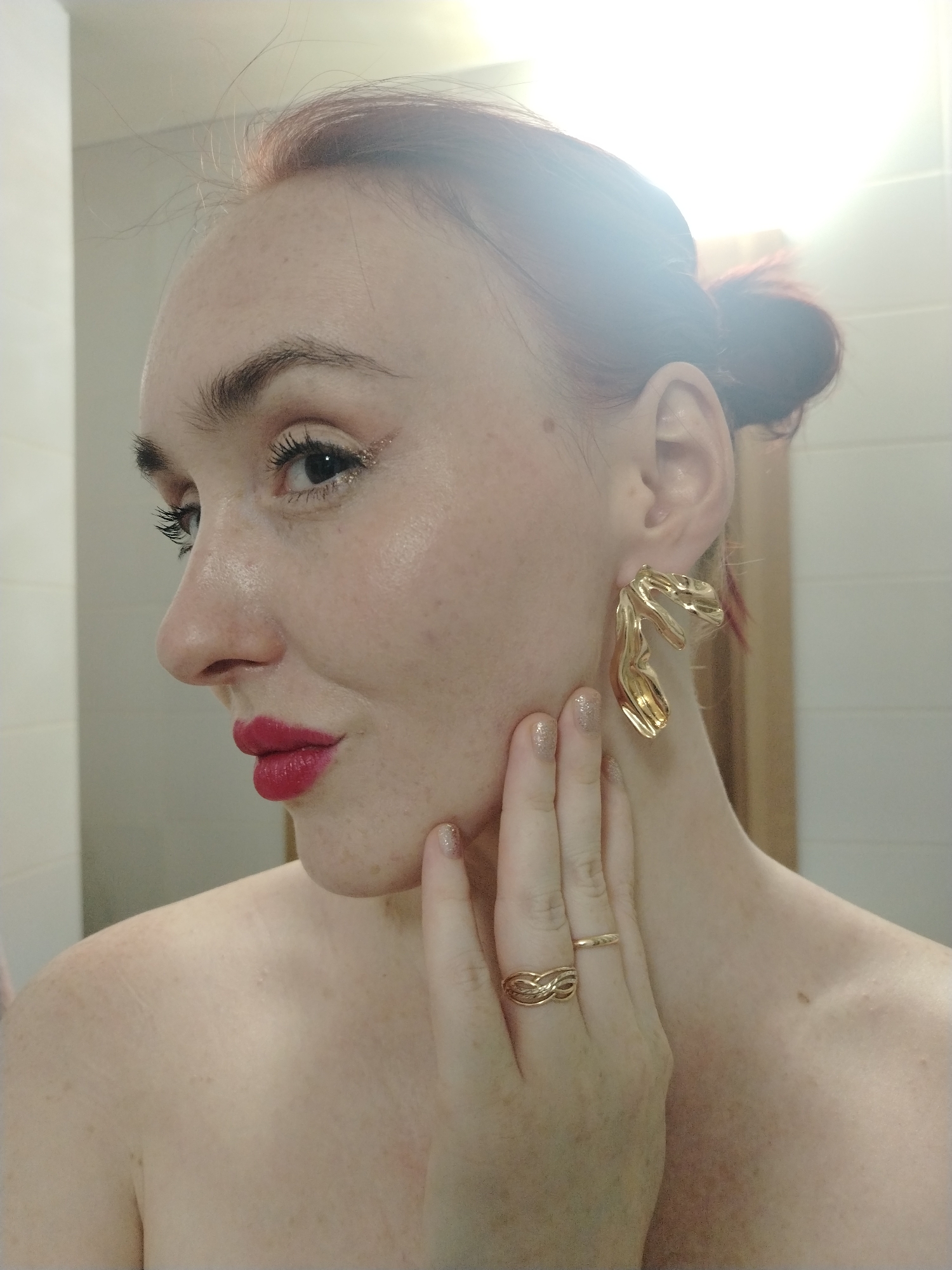 A selfie of a fair-skinned woman with freckles and red hair pulled into a bun. She is wearing gold statement earrings and two gold rings on her left hand. Her nails have a glitter polish. She is wearing red lipstick and eye makeup with gold glitter. The background appears to be a bathroom with light-colored tiles. There is no information to place the image in Izhevsk, Russia.