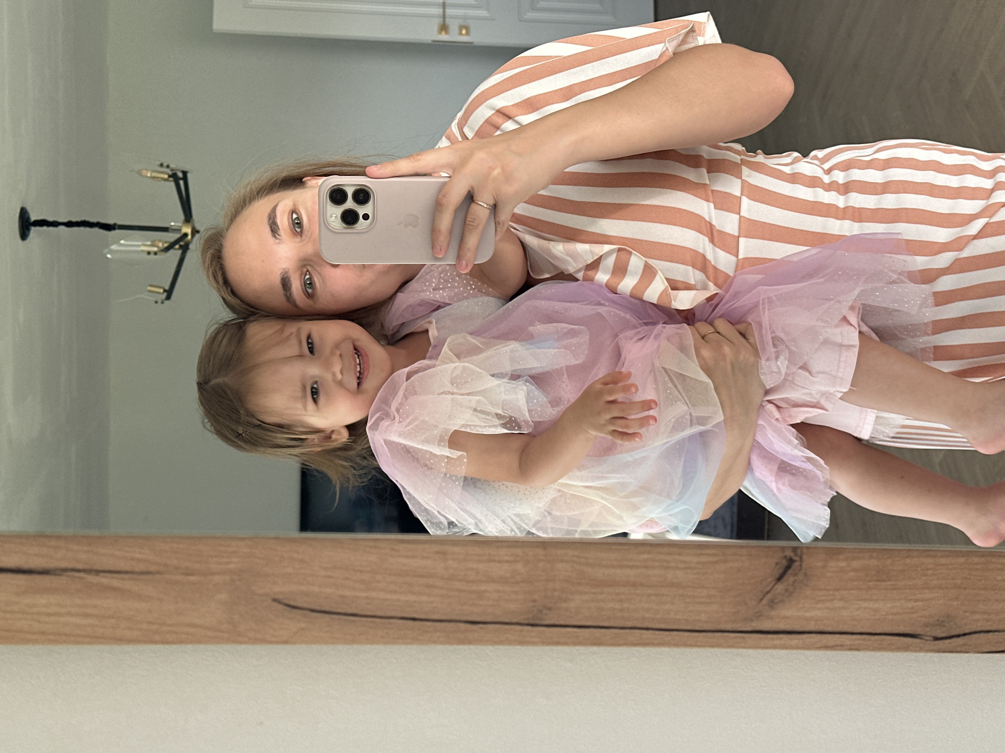 A woman stands in front of a mirror, holding a young child. The woman is wearing a striped orange and white dress and is taking a selfie with her phone. The child is wearing a pink, purple, and white sparkly dress. The background of the mirror reflects a room with a chandelier and a door. The overall scene suggests a casual moment captured at home. The exact location in the city of Kharnavy, Russia, cannot be determined from the image itself.