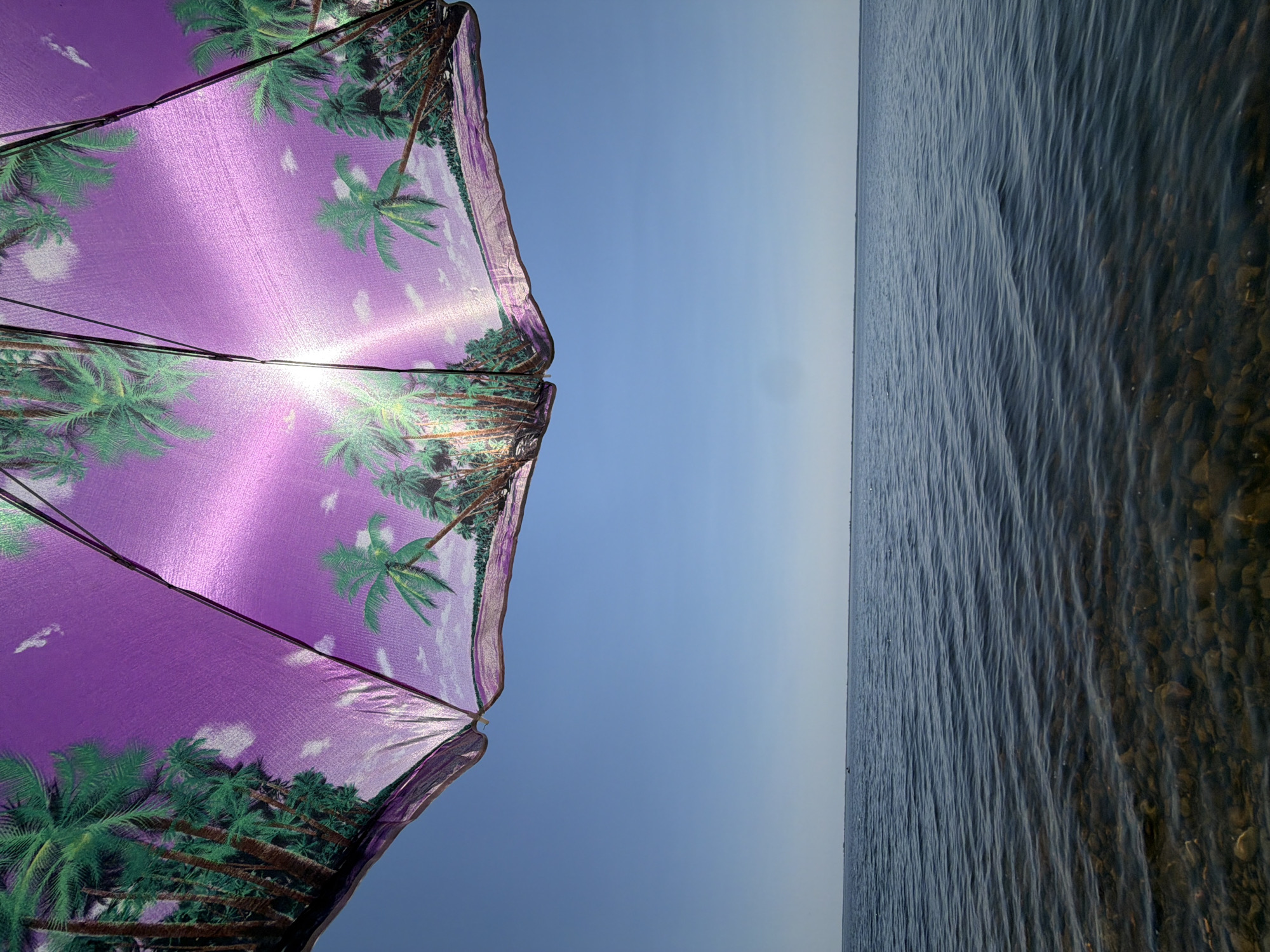 An underside view of a beach umbrella with a purple tropical scene design of palm trees against a bright blue sky. The sun shines through the umbrella, creating a lens flare. Below the umbrella is a wide expanse of calm, dark blue water with visible pebbles and rocks in the shallow foreground. The horizon line is clear and flat, with no discernible landmarks. The image is taken from a low angle, looking upwards at the umbrella and outwards towards the water.