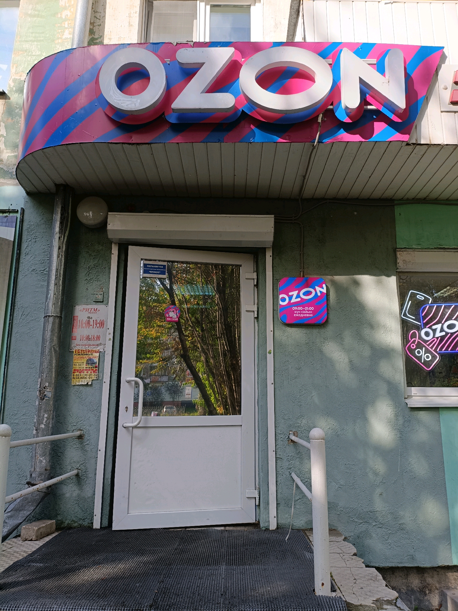 The image shows the exterior of an Ozon store located in the city of Asbest, Russia. A large, illuminated sign with the Ozon logo is prominently displayed above the entrance. The entrance itself is a white door with a glass panel, reflecting the surrounding trees and buildings. A smaller Ozon sign is mounted on the wall to the right of the door, displaying business hours. There are also various posters and stickers on the door and the wall near the entrance. The building appears to be a ground-floor establishment with a textured, greenish-blue facade. The surrounding environment includes a sidewalk with a dark mat in front of the entrance, a white railing, and some signs of wear and tear on the building and the sidewalk. There are no people visible in the image.