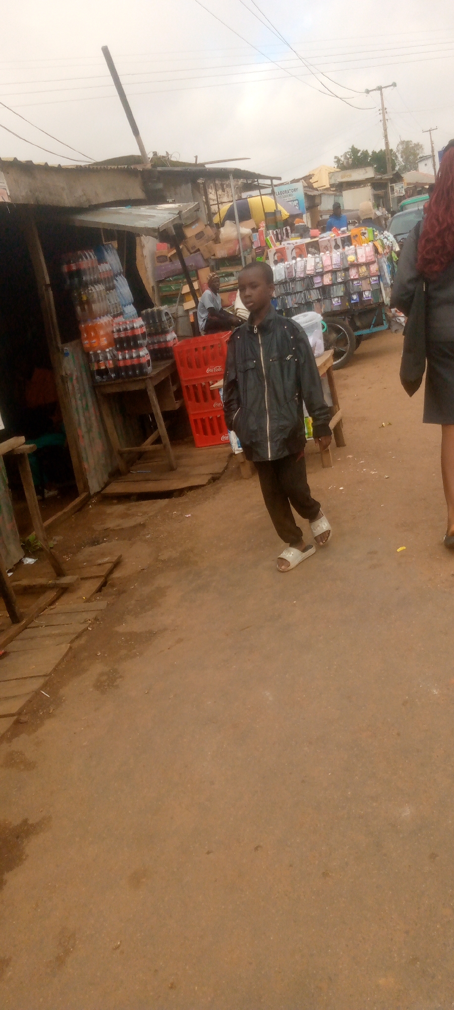 In an outdoor urban market setting, a young boy in a dark jacket and pants walks on a dirt path. He wears white slides on his feet. In the background, a vendor stall is visible, laden with various goods, including stacks of bottled drinks and what appear to be mobile phone accessories. Another person sits in the background, near the stall. The environment suggests a commercial area with a mix of stalls and open space. The sky is overcast.