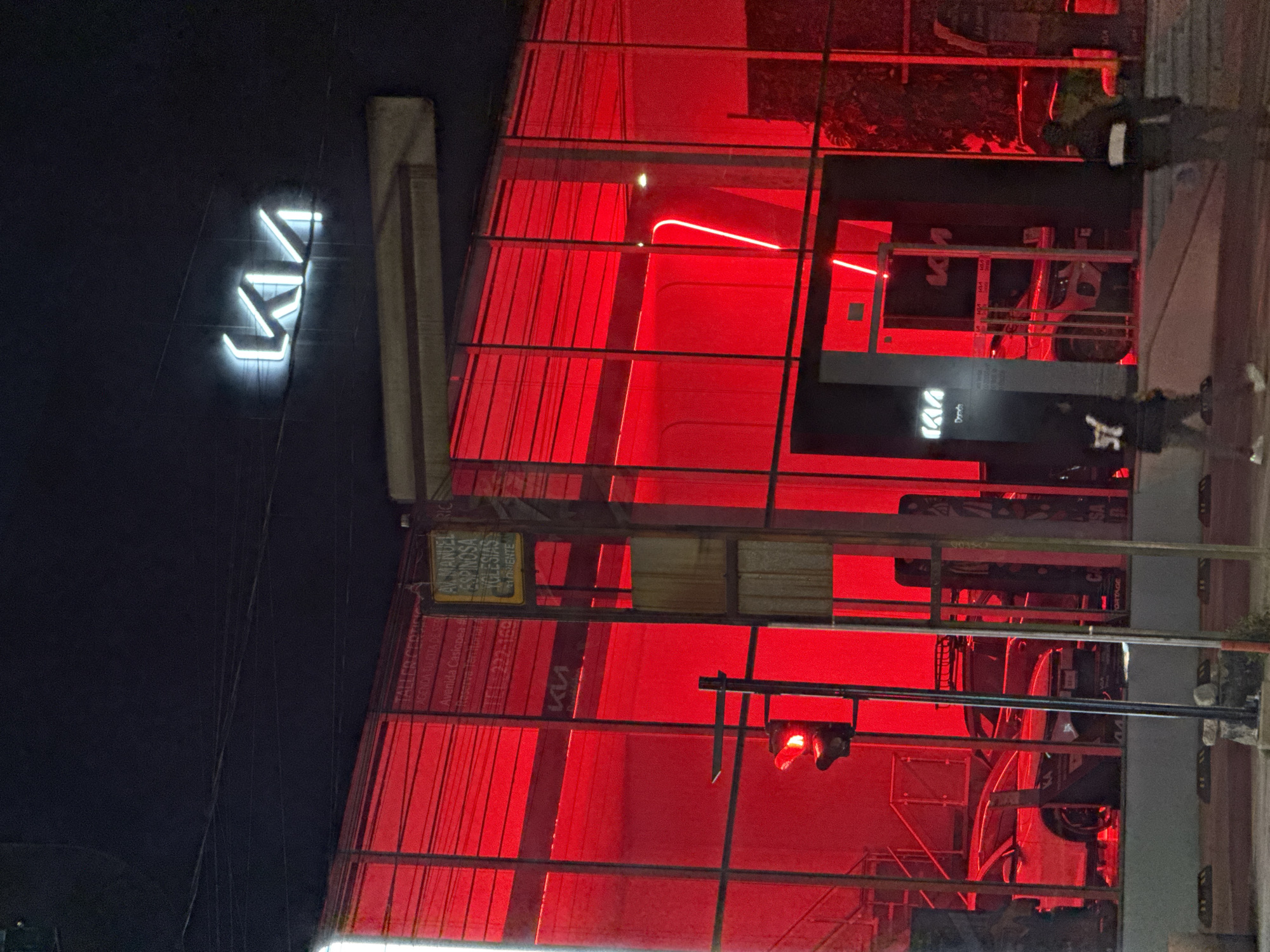 The image shows a car dealership at night in Puebla, Mexico. The dealership is illuminated with red lights. The Kia logo is visible on the building. A neon sign displays Avenida Casiopea, Av Manuel Espinosa Yglesias 21 Oriente. A pedestrian is walking on the sidewalk in front of the building.