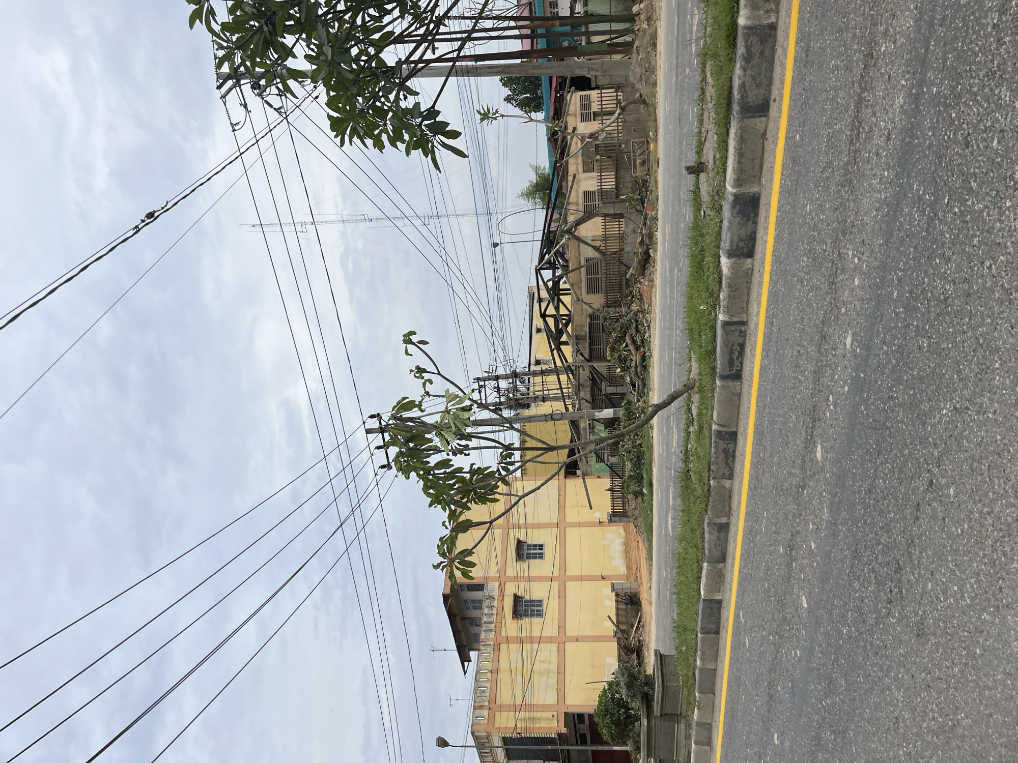 In Tanahterban, Indonesia, an image captures a street scene with a damaged building and overhead power lines under a cloudy sky. The building on the right side of the image appears to be undergoing demolition or has been partially destroyed, with its roof structure exposed and debris scattered around its base. To the left, a yellow multi-story building stands intact, featuring windows and visible structural beams. The foreground shows an asphalt road with a yellow dividing line and a curb with alternating black and grey stones, bordered by a strip of grass. Trees are visible throughout the scene, including one in the mid-ground with fallen branches and leaves on the ground, and others with green foliage to the right. Numerous power lines crisscross the sky above the buildings and road, supported by utility poles. In the background, a tall communication tower is visible. No people are present in the image.