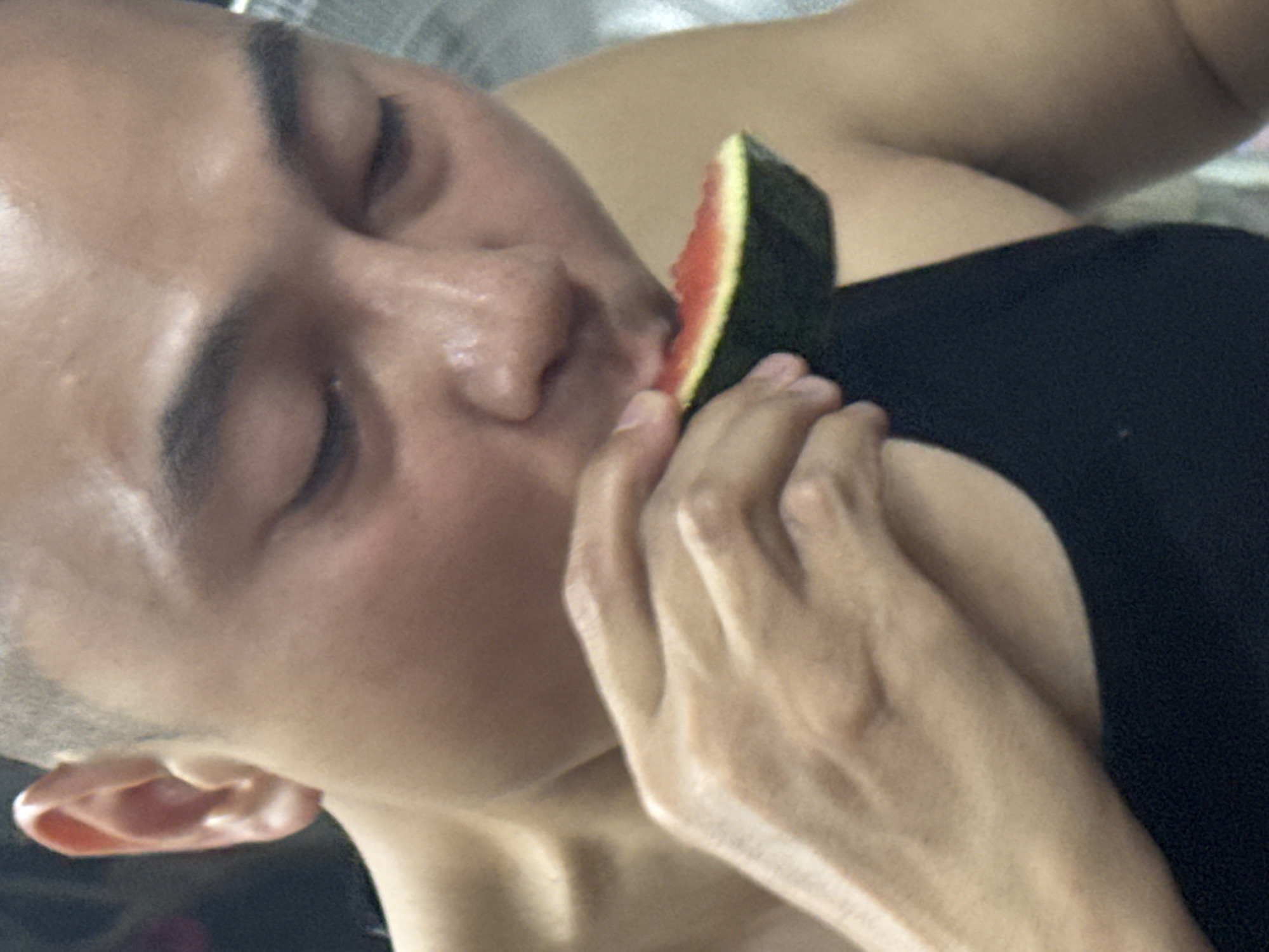 A person is holding a slice of watermelon to their mouth. The person's eyes are closed. The location is uncertain and cannot be determined from the image alone. The background appears to be a bed. The person is wearing a dark-colored shirt.