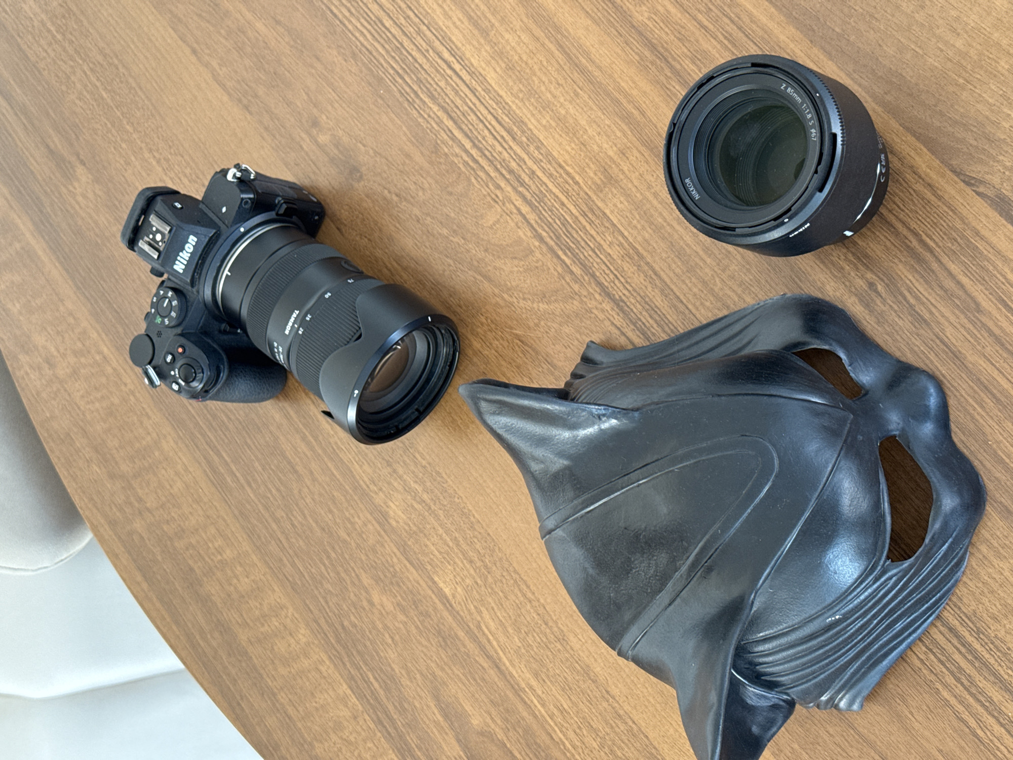 On a wooden surface, presumably a tabletop, lies a Nikon camera with a Tamron lens attached. Next to it is a Nikkor 85mm lens and a black, sculpted mask. The environment appears to be indoors. There is no evidence to suggest this image was taken in Izhevsk, Russia.