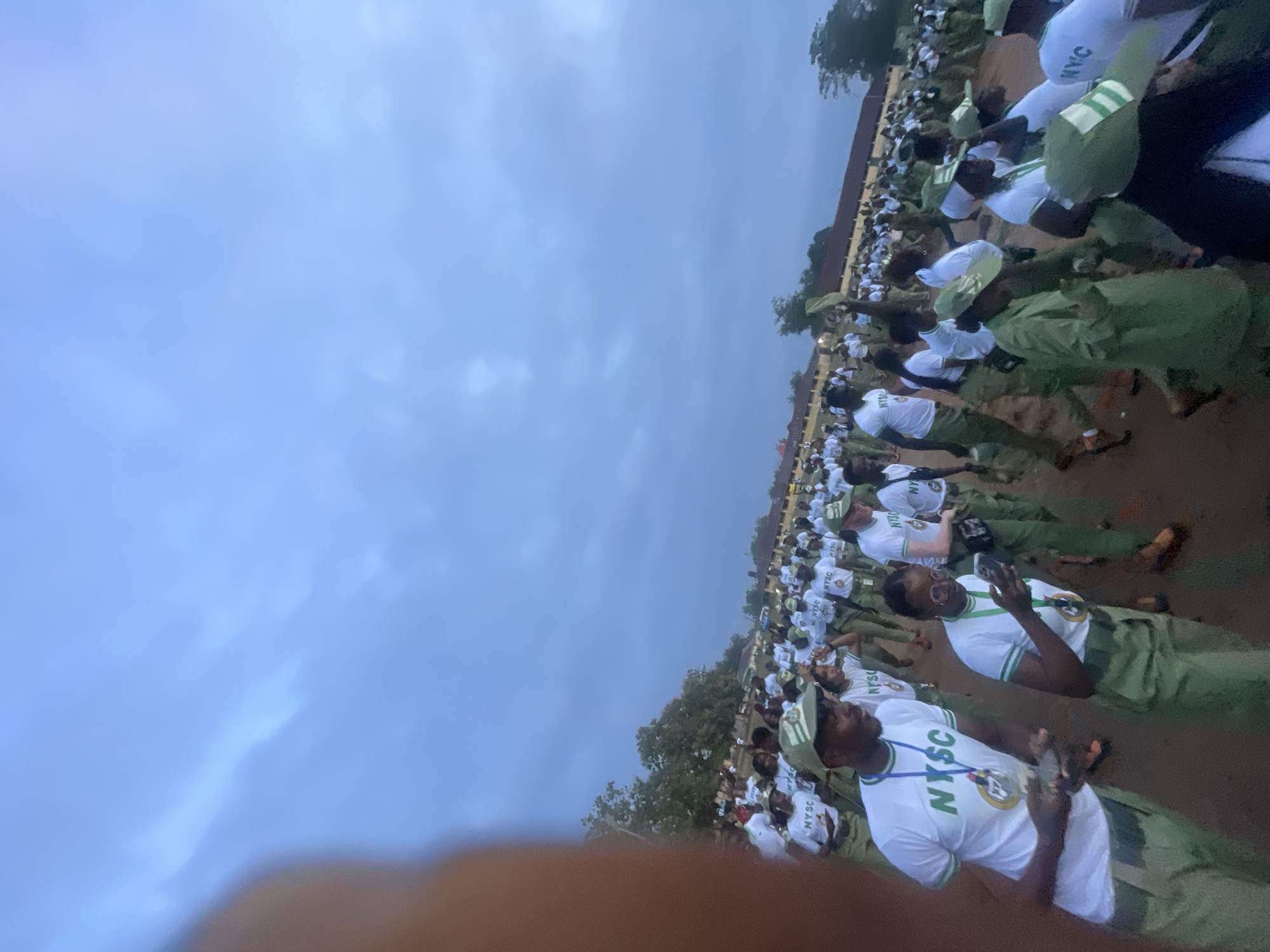 The image depicts a large gathering of young adults in what appears to be an outdoor training or orientation camp, likely at the Emure-Ekiti NYSC (National Youth Service Corps) permanent orientation camp. The individuals, predominantly dressed in white T-shirts with "NYSC" printed on them and green trousers or shorts, are participating in an activity, possibly a parade or a march. Some are holding their mobile phones, suggesting they might be documenting the event or communicating.
The environment is an open field with a dusty or sandy ground. In the background, there are buildings with reddish roofs and some trees, indicating an established camp facility. The sky is overcast with a muted, bluish-gray tone, suggesting it is either early morning or late evening. The presence of many young people in uniform points to a structured event associated with the NYSC program, which is mandatory for Nigerian graduates. The overall atmosphere suggests a communal and organized gathering.