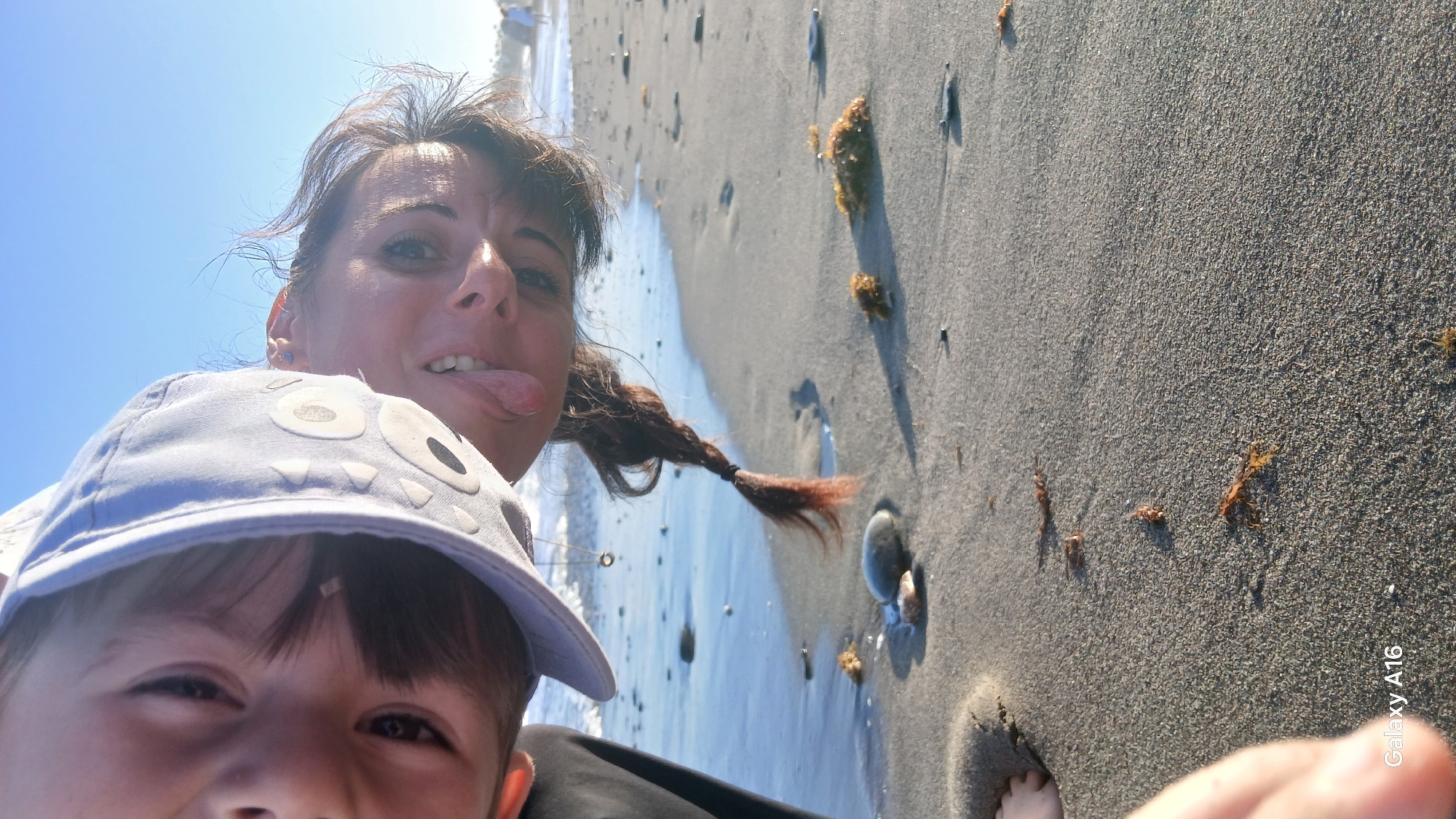 Two individuals, an adult and a child, are positioned in a selfie-style shot on a sandy beach, with the ocean and sky visible in the background. The adult, with dark hair braided into a single plait, leans forward, sticking out their tongue and smiling. They are wearing a light-colored cap with cartoonish eyes. The child, partially obscured by the adult's cap, is also looking towards the camera. The beach appears to be composed of dark sand with scattered small rocks and pieces of seaweed. The water is visible on the left side of the frame, with gentle waves reaching the shore. The sky is clear and blue. The scene suggests a casual outing at the beach. The "Galaxy A16" watermark is present in the bottom left corner, indicating the device used to capture the image. The image orientation is inverted.