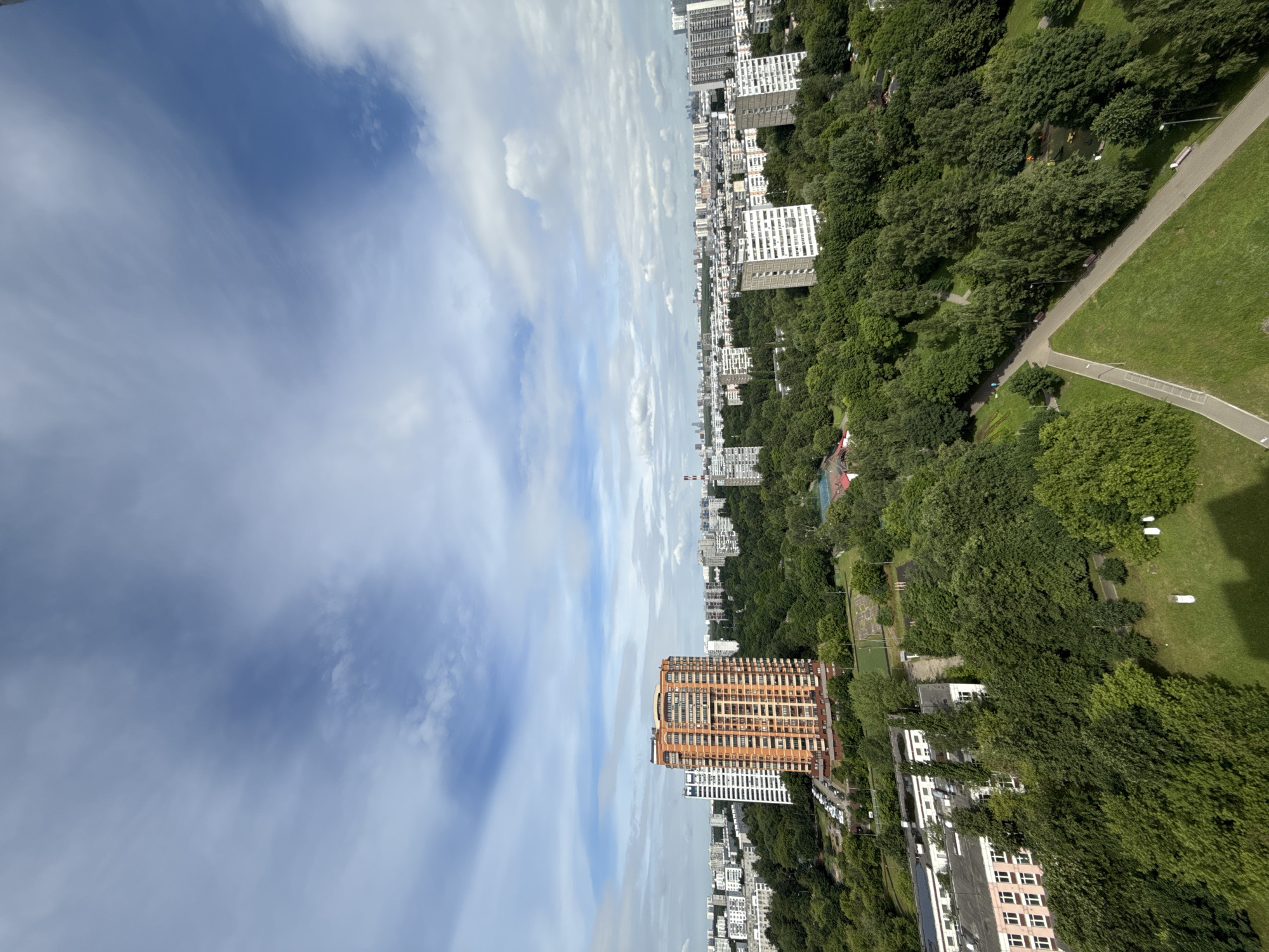 This is a high-angle, wide-angle shot of a dense urban forest bordering a residential area in Moscow, Russia. The scene is dominated by lush green trees, which fill the foreground and mid-ground, interspersed with apartment buildings of varying heights and architectural styles. In the mid-ground, a prominent tall, orange-brick apartment building stands out, surrounded by a mix of white and grey high-rise residential blocks in the background, extending towards the horizon under a partly cloudy sky. The sky itself exhibits a blend of blue patches and white, wispy clouds, suggesting fair weather. The lower left of the frame shows a school building with a pink facade and multiple windows. A network of paved pathways and green spaces, indicative of a park or recreational area, winds through the trees in the foreground and mid-ground. Cars are visible parked along a street near the orange building. The overall impression is one of a well-established urban environment with a significant presence of green space, typical of many Moscow residential districts. There are no visible people or animals in the frame, and no discernible actions or interactions.