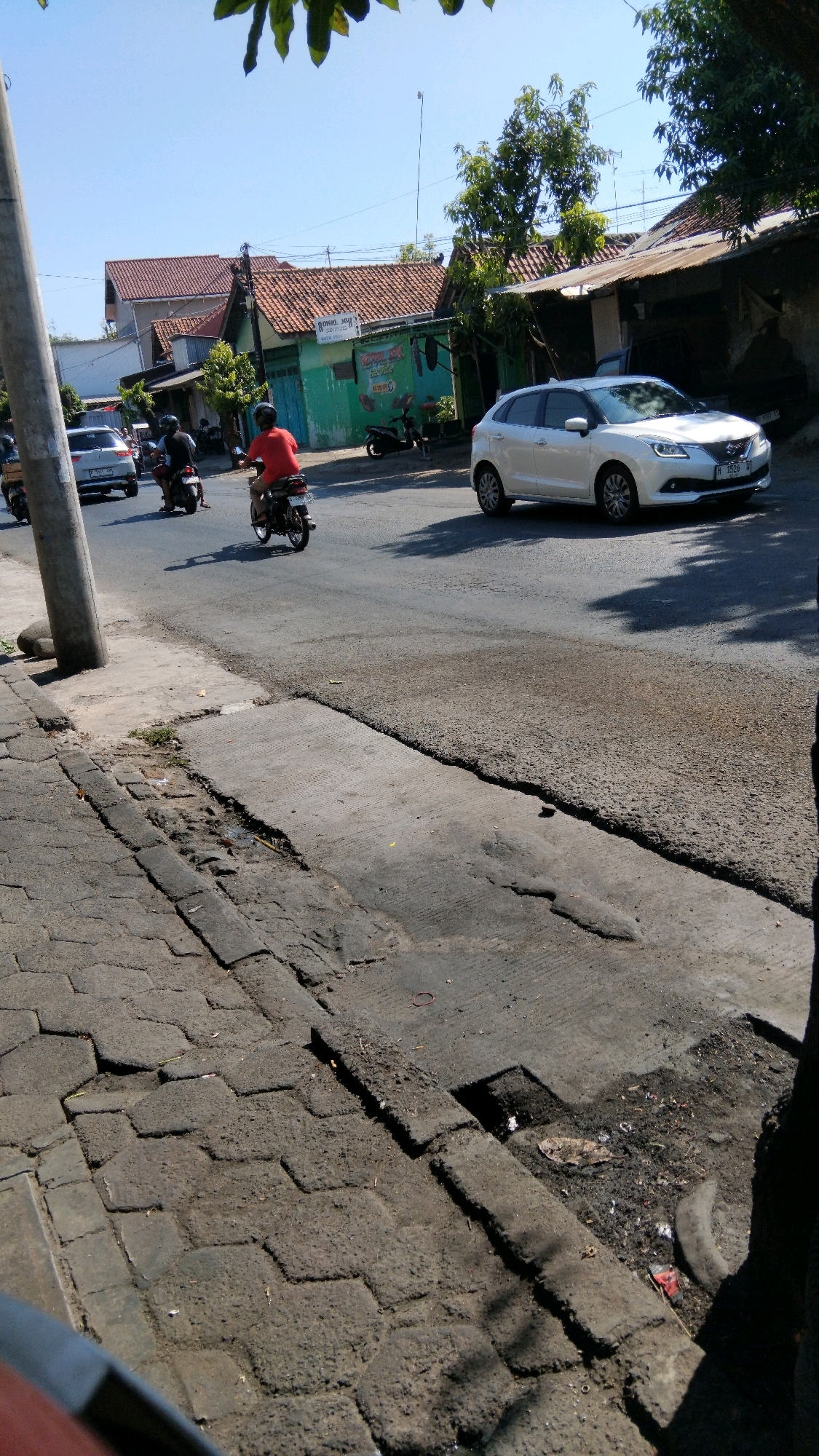 In Proyonanggan Tengah, Indonesia, on a clear, sunny day, a street scene unfolds. A white car navigates the asphalt road, its license plate partially visible. A person on a red motorcycle is riding away from the camera. Further down the road, other vehicles, including a white car and motorcycles, are present. The street is lined with buildings, some with red tile roofs and green walls, suggesting a residential or commercial area. Trees provide sporadic shade. The foreground shows a cracked concrete sidewalk and an asphalt road surface with noticeable wear and tear, indicating ongoing maintenance needs.