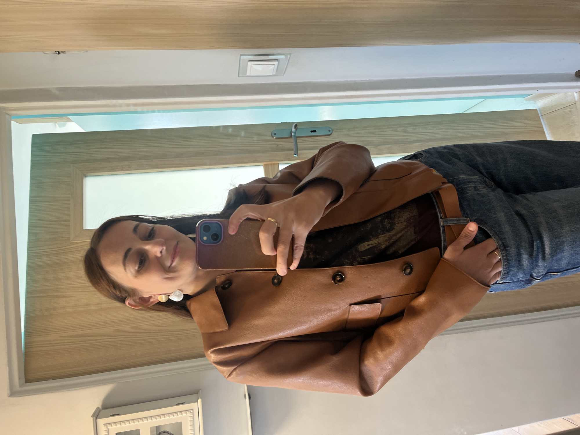 A woman stands in front of a mirror, taking a selfie. She is wearing a brown leather jacket, a dark-colored top, and blue jeans. She has on earrings and a ring on her left hand. The background appears to be a home interior, with wooden doors and a light-colored wall.