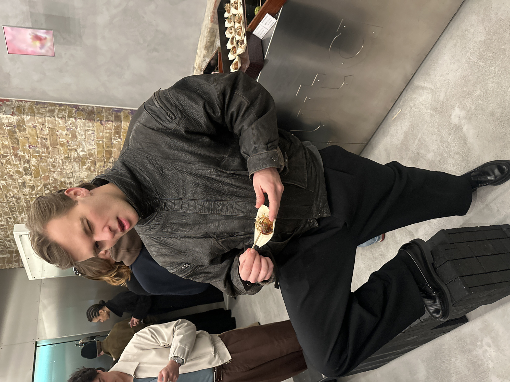An indoor event in the City of London, United Kingdom, depicts a social gathering. In the foreground, a male with light complexion, styled brown hair, and an earring in his left ear wears a dark textured leather bomber jacket, a grey shirt, black pleated trousers, and black loafers. He stands with his left foot resting on a black modular block structure, leaning against a silver-toned metallic counter with embossed letters, partially spelling "UTC". He holds a single takoyaki (Japanese octopus ball) in a small wooden boat-shaped dish with a wooden pick, while winking with his right eye and his mouth slightly ajar.
The metallic counter serves as a display, featuring additional takoyaki in similar wooden dishes arranged on dark wooden risers. A small white label with black text is positioned near the food. In the background, several individuals are present, including a male in a cream jacket and brown trousers checking his phone, and others with braided hair and a dark beanie. The environment comprises an exposed brick wall on the left, a plain grey wall on the right with a mounted framed artwork depicting a pink and yellow abstract form, and a light grey concrete floor.