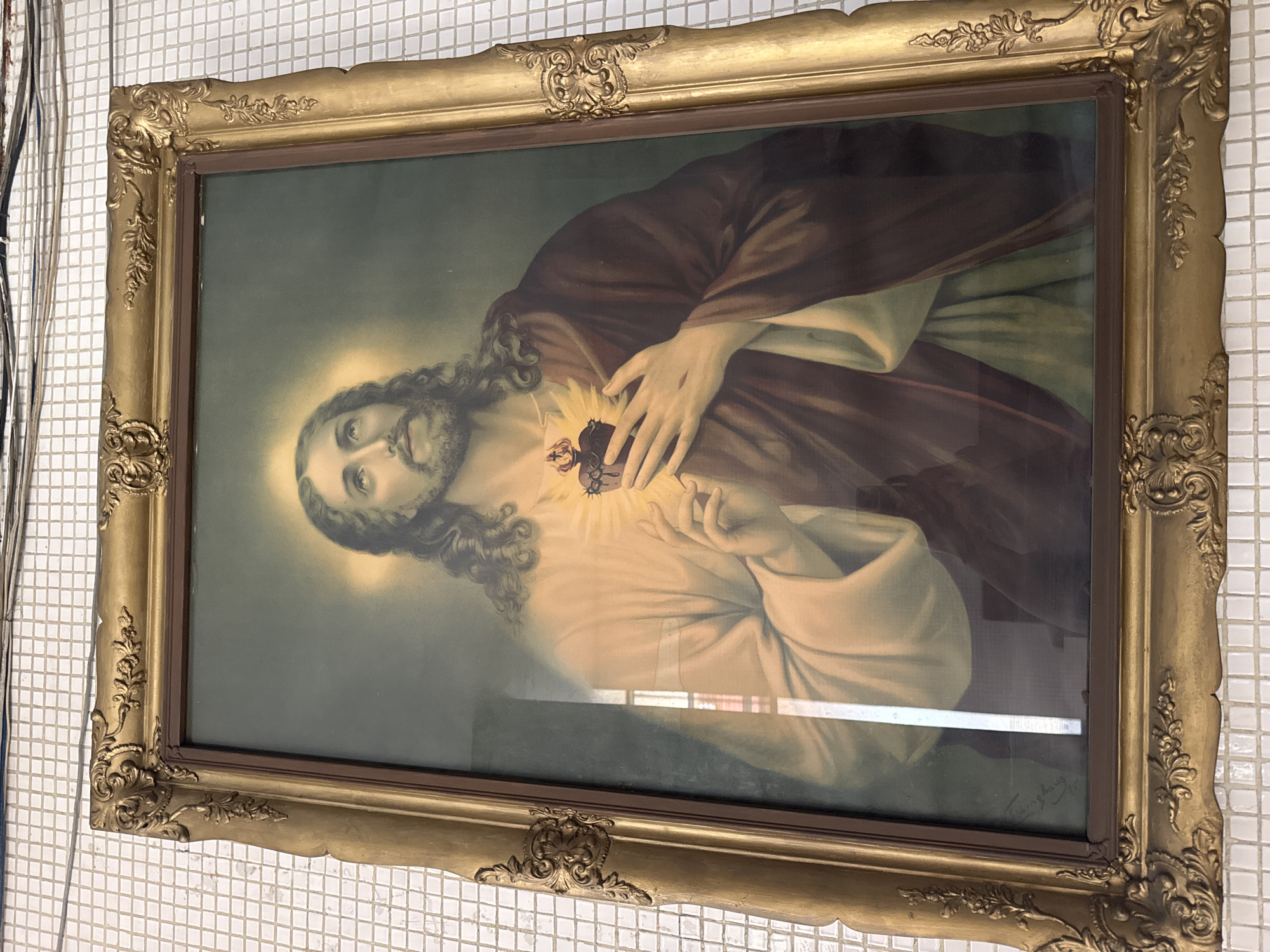 This is a framed painting of the Sacred Heart of Jesus. The subject, Jesus, is depicted with long, wavy brown hair and a beard. His eyes are open and looking slightly upwards, with an expression of gentle sorrow or contemplation. He is wearing a dark reddish-brown robe draped over his left shoulder, revealing a lighter garment underneath. His right hand is placed over his chest, pointing towards his exposed heart, which is depicted as flaming and crowned with thorns, with a white lily emerging from it. His left hand rests gently on the robe. A halo of light surrounds his head.
The painting is framed in an ornate, gilded frame with intricate scrollwork and floral motifs, particularly at the corners. The background of the painting is a dark, muted green, creating a somber atmosphere. A faint signature is visible in the lower right corner of the painting.
The painting is hanging on a wall covered in small, white square tiles. The lighting in the room appears to be artificial, as indicated by the reflections on the glass covering the painting, which show some light fixtures. There are also some electrical wires visible on the left side of the image, suggesting an indoor setting. The image itself is oriented vertically, indicating the painting is hung in portrait orientation. There is no visible indication of time of day or weather. The location context is Maracay, Venezuela.