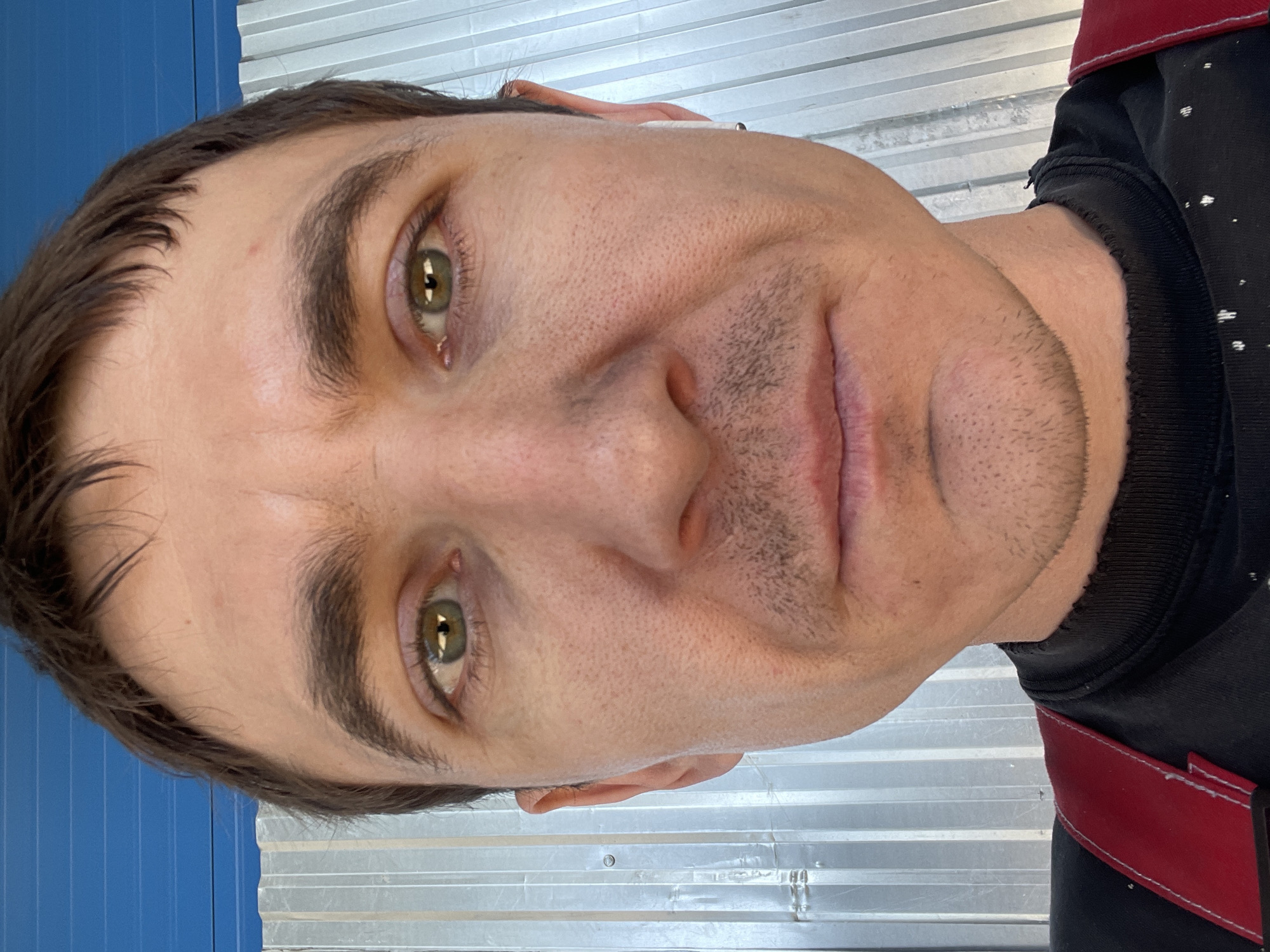 The image is a close-up, front-facing selfie of a man with brown hair, green eyes, and light stubble on his face. He is wearing a black t-shirt with red suspenders. The background consists of a corrugated metal wall and a blue surface. The lighting is natural, casting shadows on the man's face. The photo appears to have been taken outdoors, possibly in a rural or industrial area. The man's expression is neutral. No specific location in Sokury, Russia, can be identified from the visual information provided.