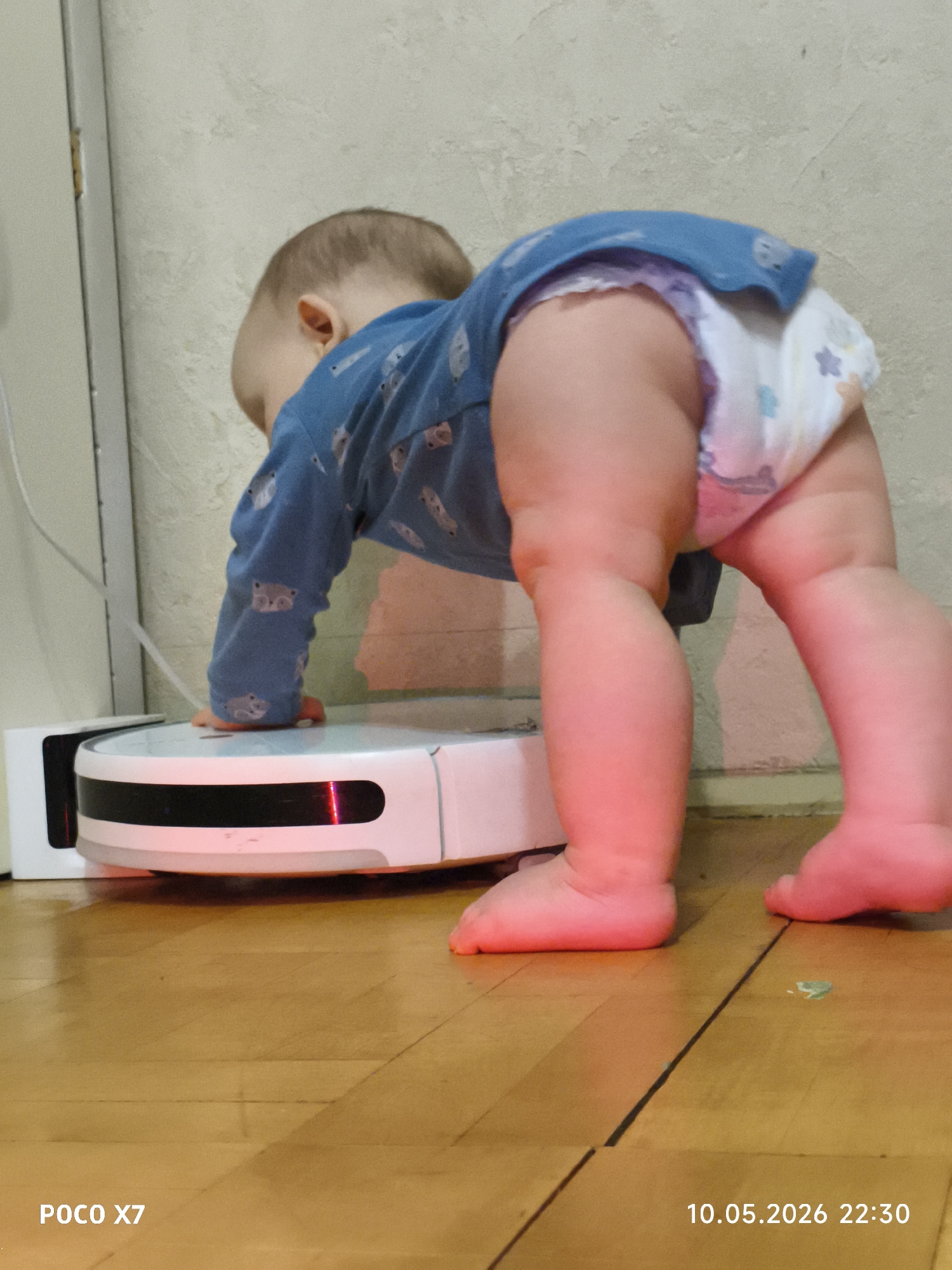 A baby is on all fours, leaning on a white robot vacuum cleaner with a red light. The baby is wearing a blue shirt and a diaper. The scene is set indoors on a wooden floor near a white wall and a white door frame. The timestamp in the bottom right corner reads "10.05.2026 22:30" and "POCO X7" is on the bottom left. The city of Chaikovsky, Russia is not directly indicated in the image, therefore its presence cannot be confirmed.