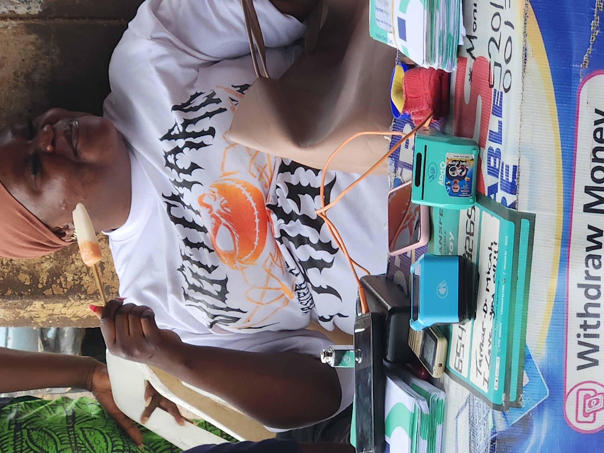 The image depicts an outdoor scene, likely a street market, with a woman in the foreground holding a food item on a skewer. She is wearing a white t-shirt with a graphic design and a brown headscarf. In front of her, a table is laden with various items, including stacked papers, cell phones, payment terminals, and a beige handbag. The background is indistinct, suggesting a busy urban environment. While the specific location within Jos, Nigeria, cannot be definitively determined from the image alone, the presence of local currency denominations on some of the papers and the general atmosphere are consistent with a Nigerian setting. The overall impression is that of a transaction or a moment of pause during a commercial activity.