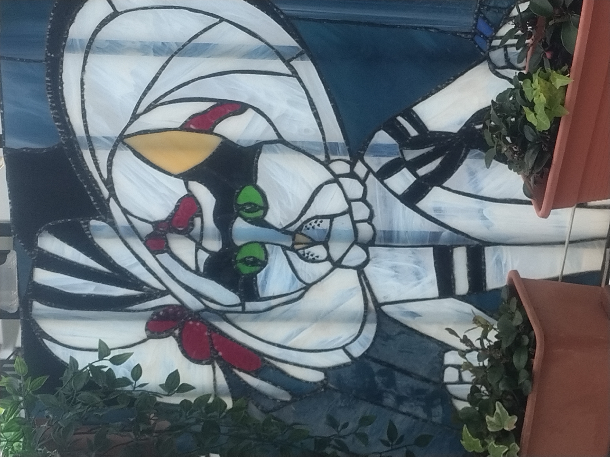 A stained-glass panel depicting a stylized cat is installed vertically on a wall outdoors. The panel features a white cat with black accents, including stripes on its body, set against a background of blue and white glass. The cat has green eyes, a yellow nose, and red details. Planter boxes containing leafy plants flank the panel, adding to the scene. The city is Luxembourg, Luxembourg.