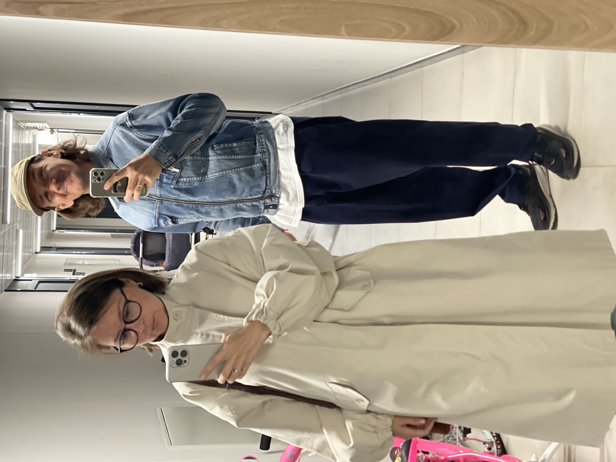 A vertically oriented medium shot depicts two individuals reflected in a mirror. The upper individual, a male, wears a tan baseball cap, blue denim jacket, white t-shirt, navy slacks, and dark athletic shoes. He holds a smartphone in his hand, presumably taking the picture. The lower individual, a female, wears glasses and a light-colored trench coat. She also holds a smartphone. The setting appears to be a hallway, possibly in a commercial or residential building. In the lower left corner, a pink child's bicycle is partially visible. The location is stated to be Izhevsk, Russia. No specific landmarks or contextual clues confirm the precise location within the city.