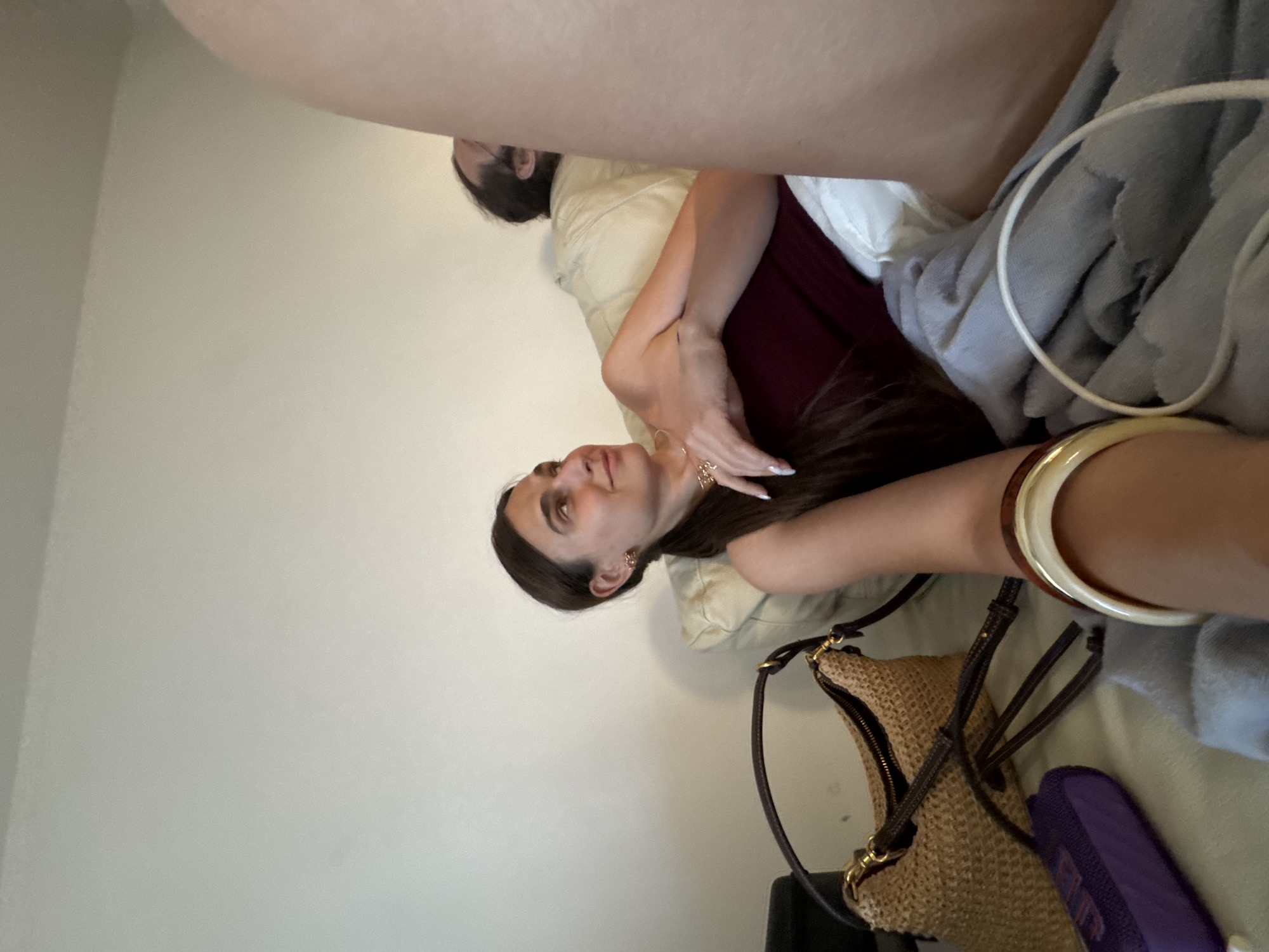 An outdoor, natural-light photograph captures a woman lounging on a couch, her body angled towards the right of the frame, with her gaze directed upwards and to the right. She has long, dark hair tied back in a ponytail and is wearing a maroon tank top and white shorts. A straw shoulder bag with dark leather straps rests on the couch to her left, and a purple object is partially visible beneath it. A light blue blanket covers part of the couch, with a white cord resting on top. A man is visible in the background, reclining on the couch to the woman's right, his head turned away from the camera. The background consists of a plain, light-colored wall. The image does not contain enough visual information to determine the specific location within Budva, Montenegro.