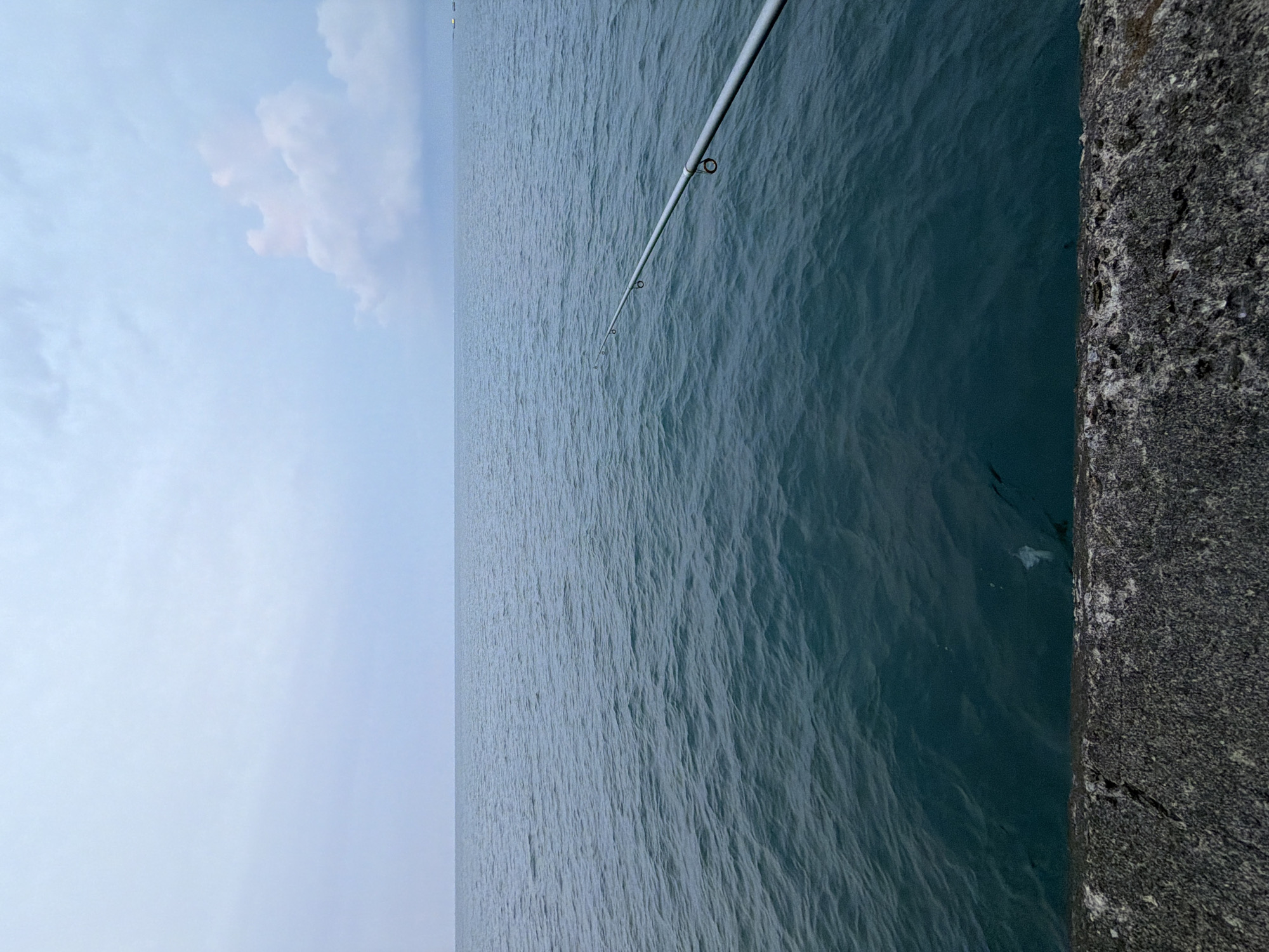 The image depicts a serene seascape, likely taken from a pier or jetty in Lhokseumawe, Indonesia. The foreground is dominated by a weathered, grey concrete structure, possibly part of a harbor or breakwater. Beyond this structure, the vast expanse of the ocean stretches to the horizon, its surface a deep, tranquil blue-green, disturbed by gentle ripples. A fishing rod, its metallic shaft and guides clearly visible, extends diagonally across the water, suggesting an angler is present, though unseen. The sky above is overcast with a soft, diffused light, hinting at an impending sunset or a cloudy day. A solitary, large cloud formation, tinged with a subtle pinkish hue, breaks the uniformity of the grey sky on the right side of the frame. The overall impression is one of calm and quietude, characteristic of a peaceful fishing spot.