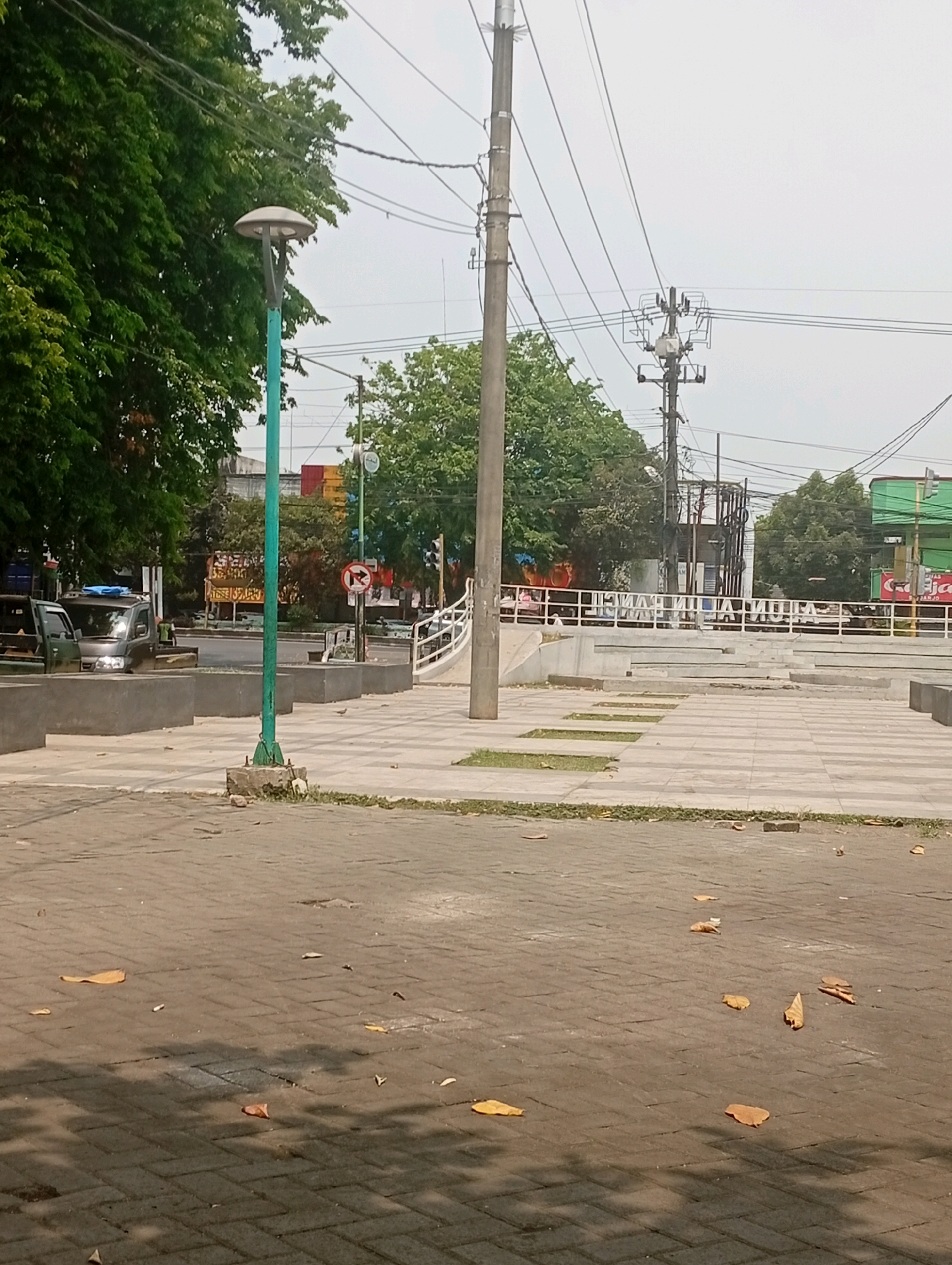 The image shows an outdoor scene with a paved walkway in the foreground, scattered with fallen leaves. A green lamppost stands to the left, with a rounded lamp fixture. Beyond the walkway, there is a street with two parked vehicles, one of which is a silver truck with a blue tarp covering its bed. Trees with lush green foliage line the background, and utility poles with numerous wires crisscross the sky. In the distance, there are buildings, including one with a green facade and a sign that appears to be in Indonesian. A traffic signal and a no-turn sign are visible near the street. The overall impression is that of a streetscape in a moderately urban environment, possibly in Bangil, Indonesia, given the architectural style and signage. The sky is overcast, suggesting a cloudy day.
