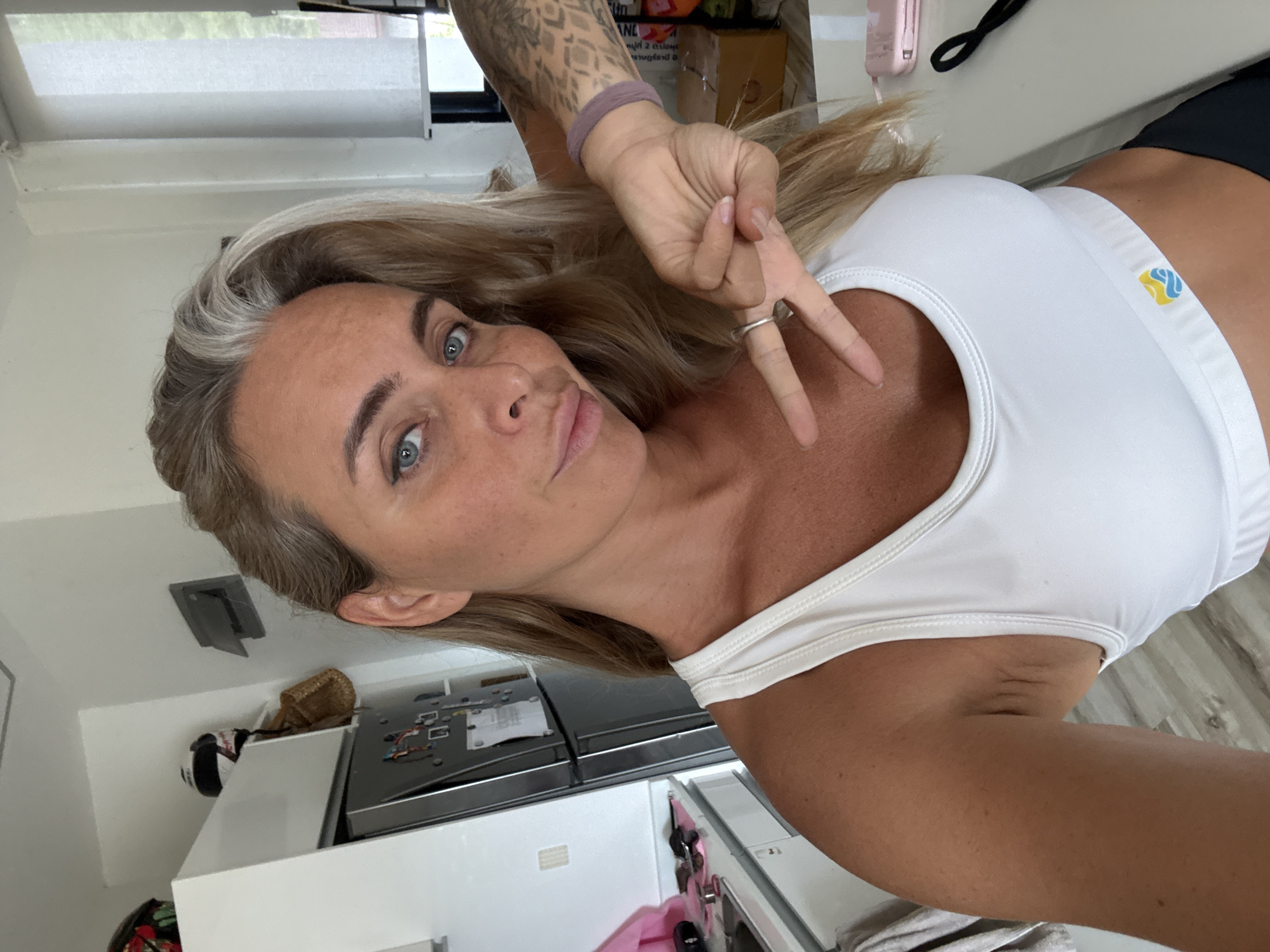 A woman with blonde hair and blue eyes is taking a selfie in what appears to be a kitchen. She is wearing a white sports bra. Her left hand is raised, forming a peace sign. She has a tattoo on her right arm. In the background, a refrigerator, cabinets, and a window with a roller blind are visible. There are also some boxes and other kitchen items in the background. The scene appears to be indoors, in a residential setting, possibly in the city of Ko Samui District, Thailand, given the lighting and general ambiance.