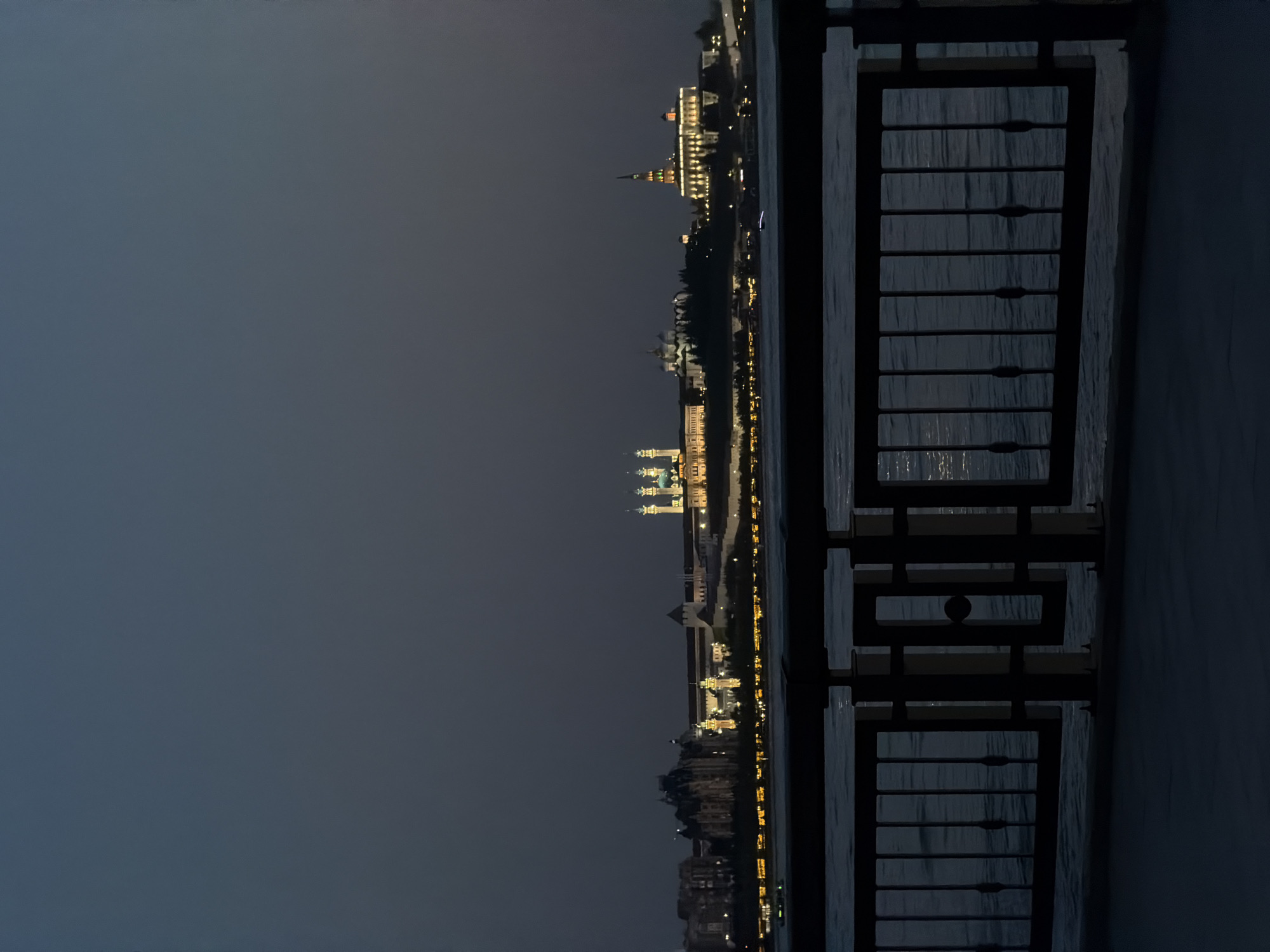 At night, a view of the city of Kazan, Russia, is seen through a dark wooden structure, likely a fence or railing. The sky is dark and the city is illuminated with lights. The light is reflected in the water below. The scene suggests an observation point from a bridge or waterfront area. The absence of people or significant activity emphasizes the focus on the cityscape and the night ambiance.